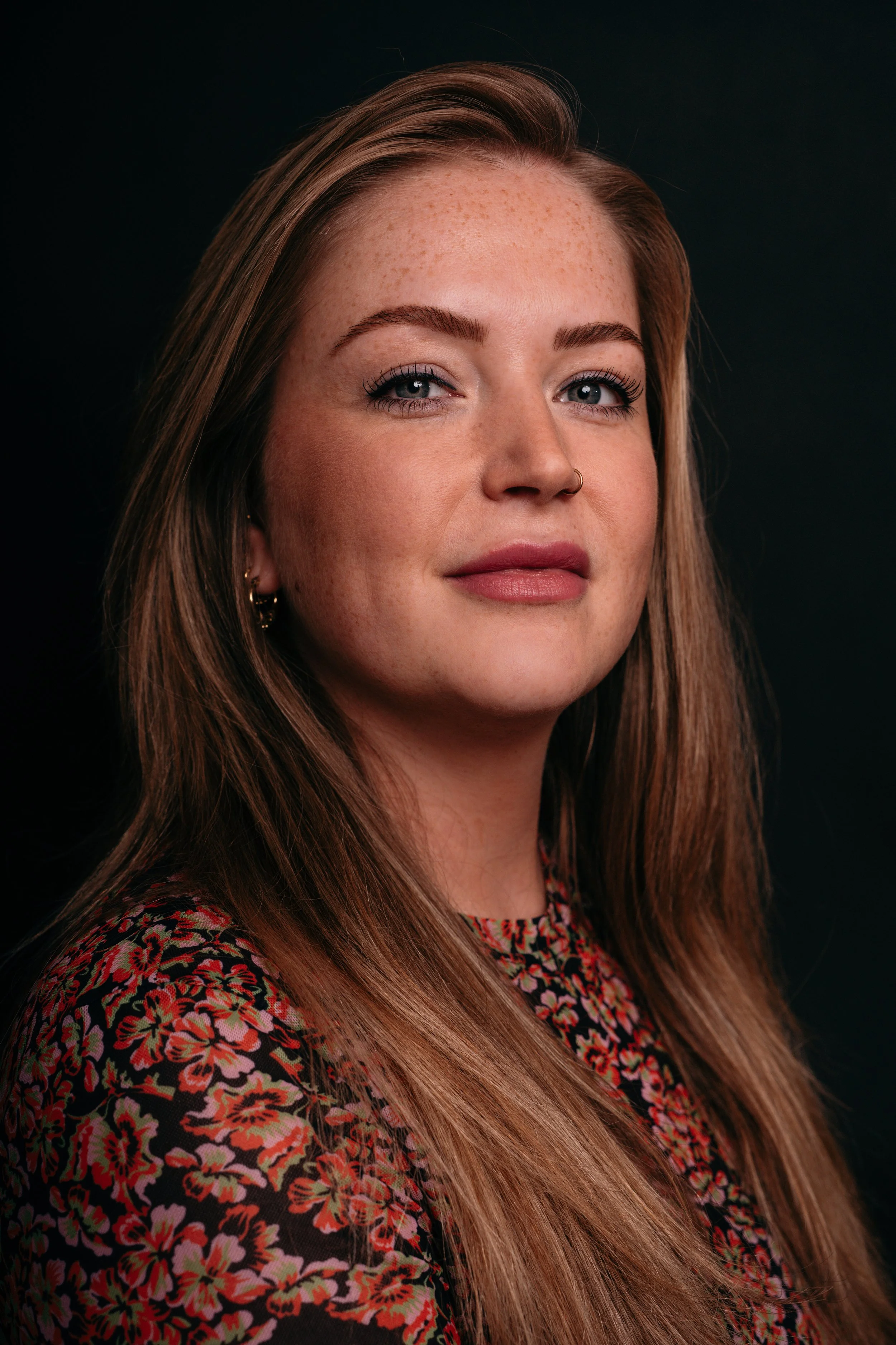 Portret van een jonge vrouw met lang, rood haar en een bloemvormige top, glimlachend in een studio met een donkere achtergrond.