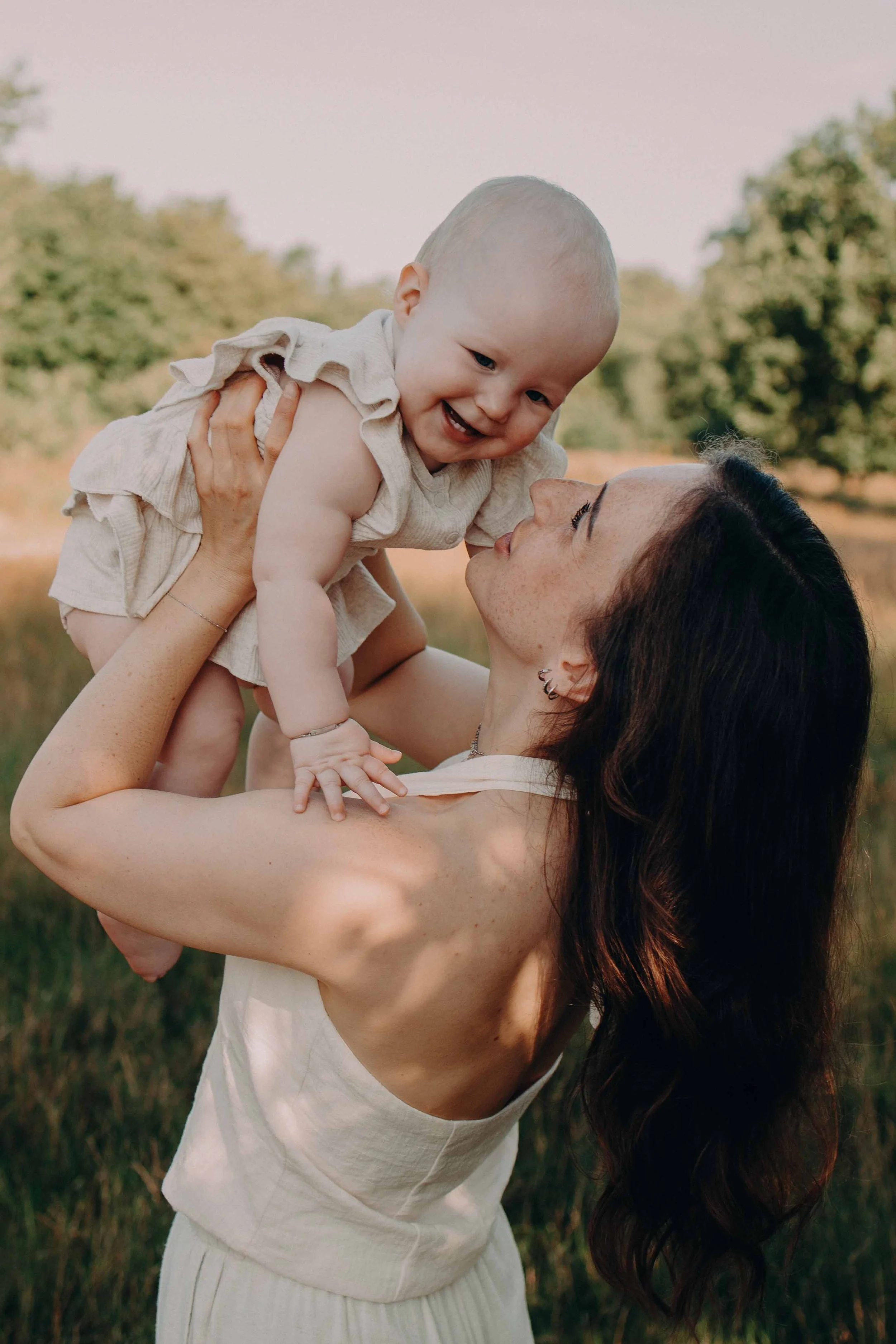 Een vrouw houdt een lachende baby omhoog in een park met groene bomen op de achtergrond.