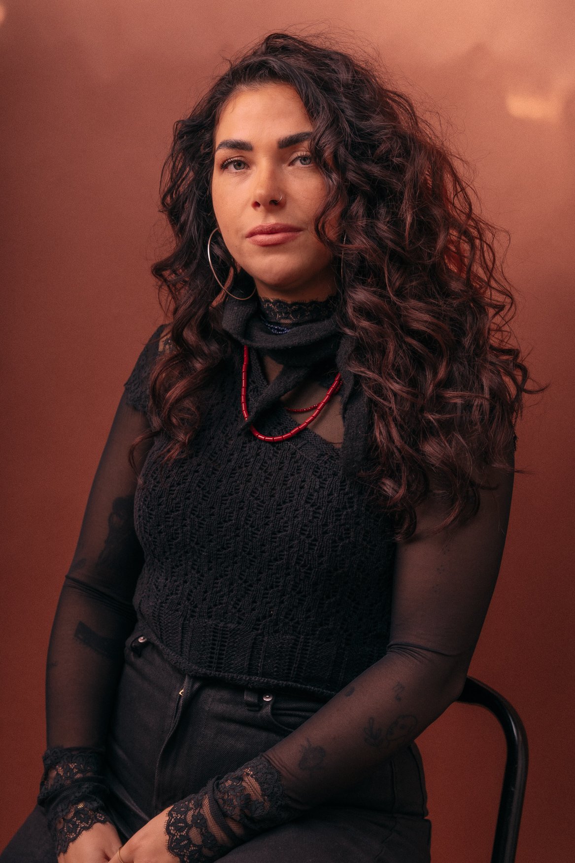 Portret van een vrouw met lange, krullende, donkere haar, gekleed in zwart met sieraden, zittend voor een effen bruin achtergrond.