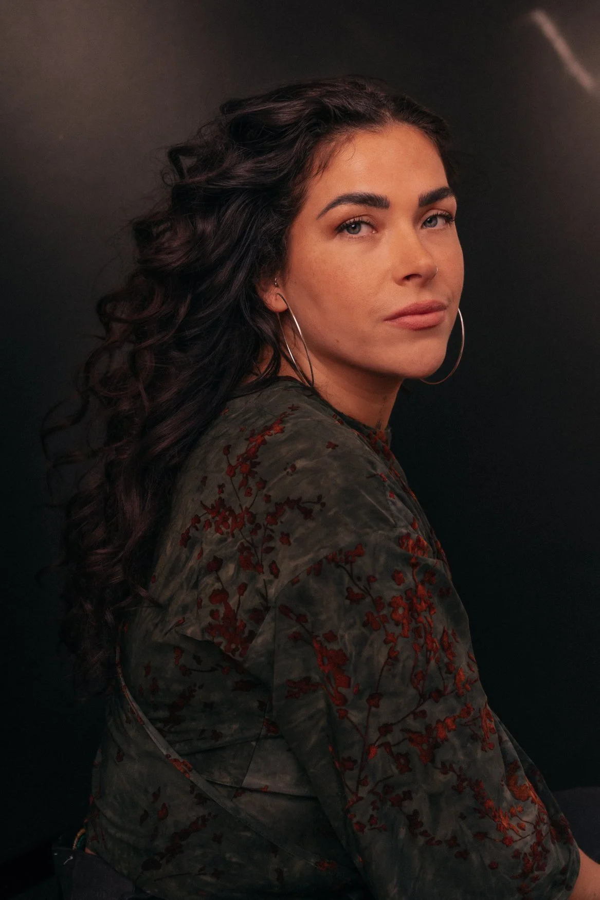 Portret van een vrouw met lange, krullende haar, grote oorbellen, een donkere blouse met bloemendessin, zittend tegen een zwarte achtergrond.