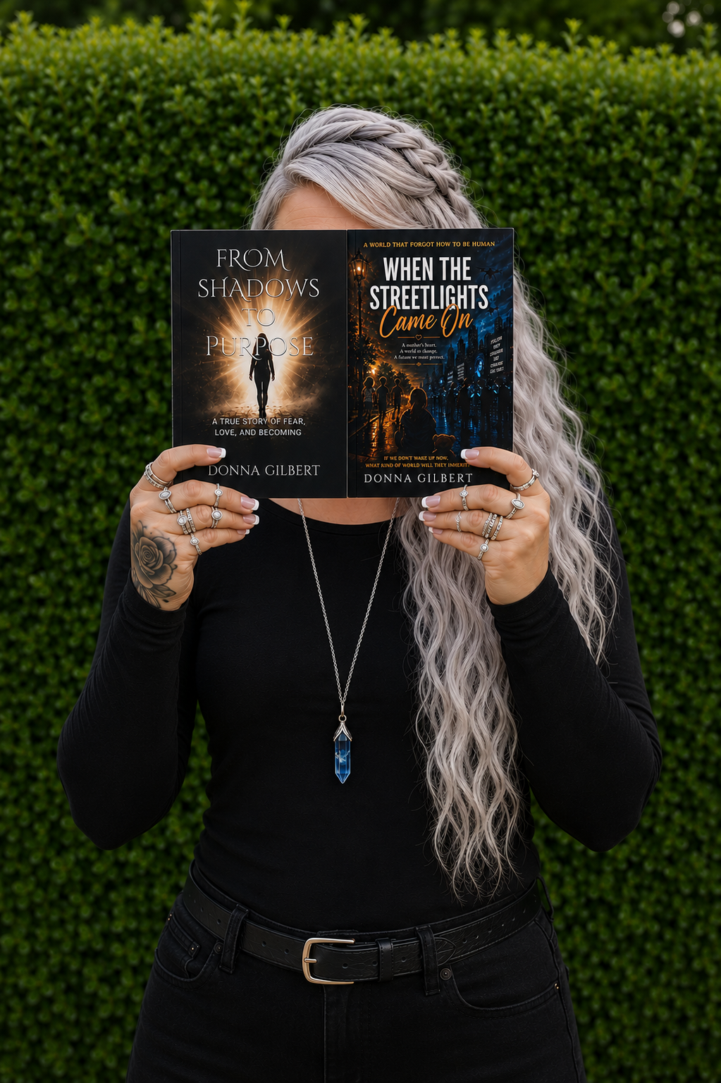 A woman with long, curly, grayish hair and multiple rings on her fingers is holding two books in front of her face, obscuring it. She is standing outdoors in front of a green hedge. She is wearing a black long-sleeve shirt, black pants, a black belt, and a long necklace with a blue crystal pendant. The books she is holding are titled 'From Shadows to Purpose' and 'When the Streetlights Came On', both authored by Donna Gilbert.