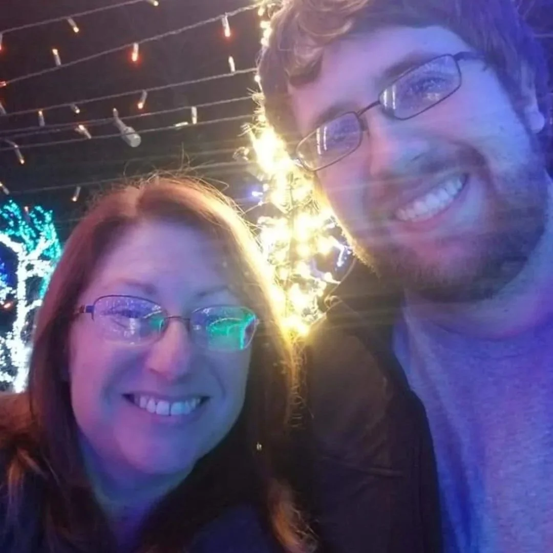 A smiling woman and man taking a selfie at night with Christmas lights and decorations in the background.