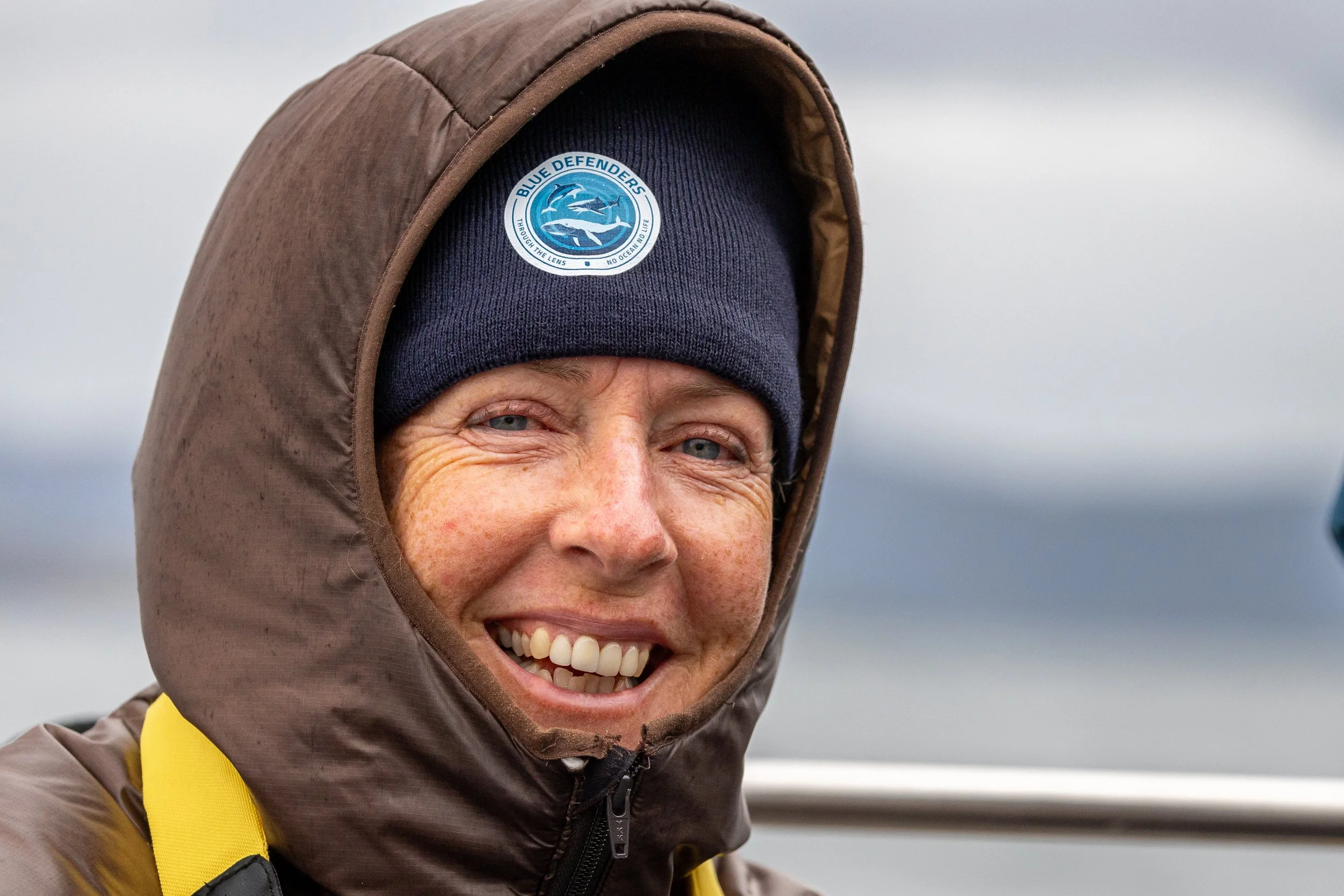 Close-up of a smiling woman in outdoor gear, wearing a dark blue beanie with a 'Blue Defenders' logo and a brown hooded jacket, with a blurred outdoor background.