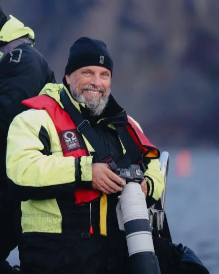 A smiling man in outdoor gear holding a camera with a telephoto lens.