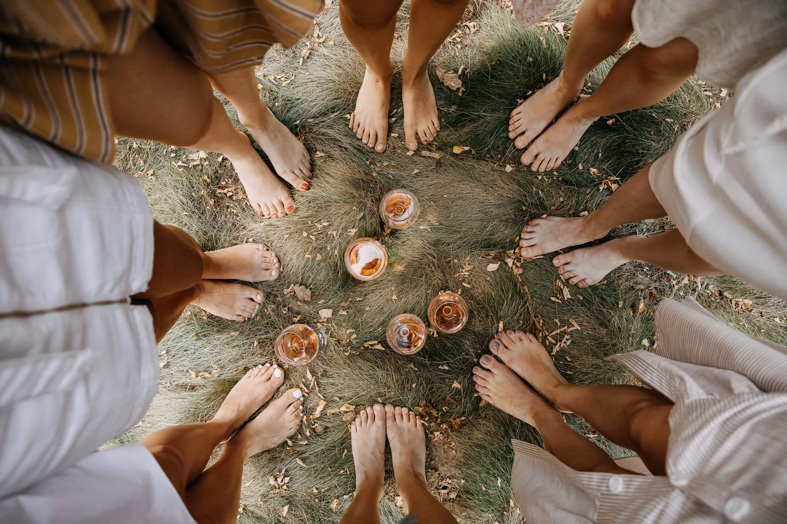 Six women standing barefoot in a circle, looking down at four glasses of rosé wine placed on dry grass with fallen leaves.