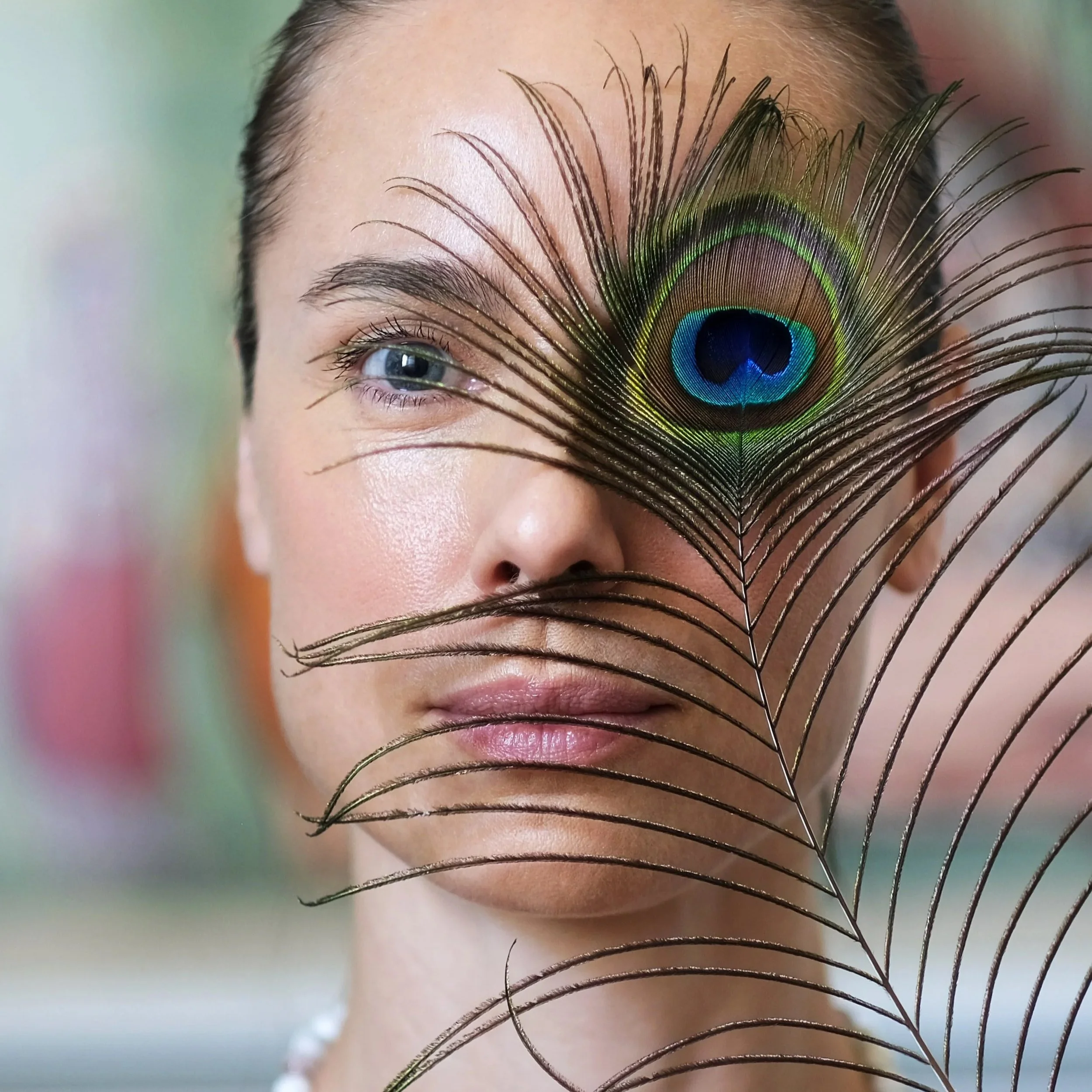 A woman with a peacock feather covering part of her face, the feather obscures one eye and her nose, but her other eye and lips are visible.