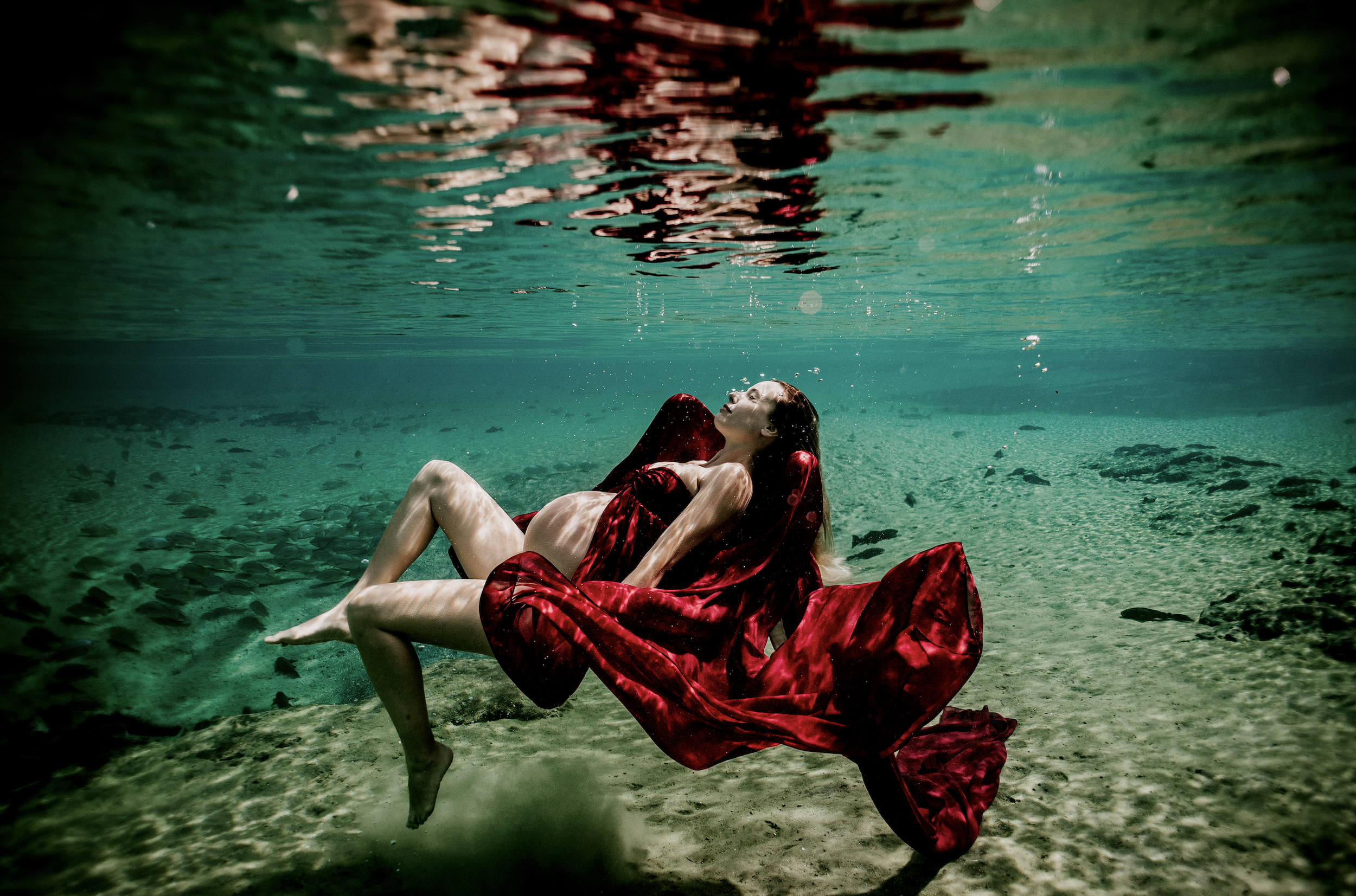 A pregnant woman wearing a red dress reclining underwater in a clear river or lake, with light filtering through the water and rocks visible on the lakebed.
