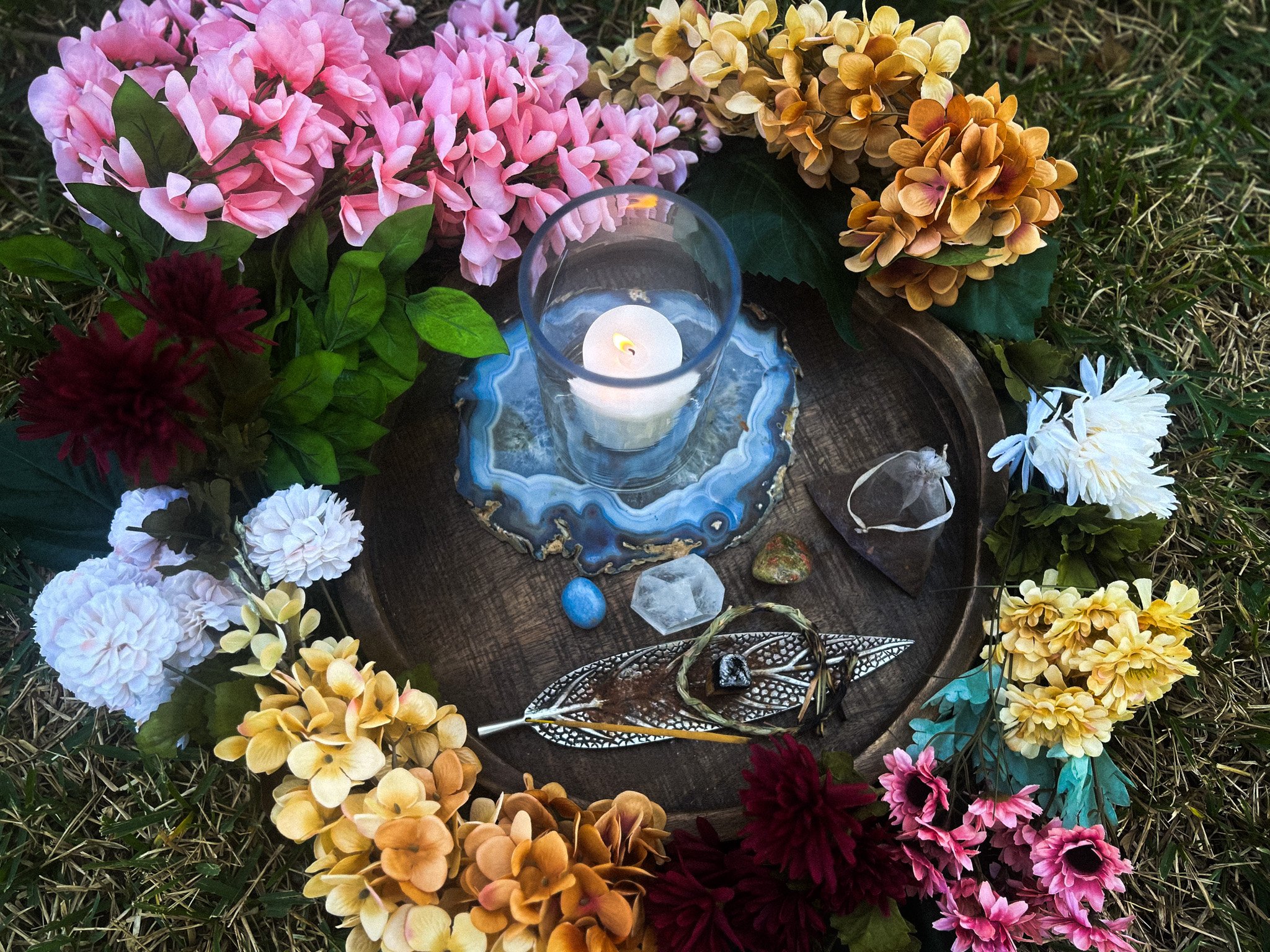 A circular arrangement of colorful flowers surrounding a wooden tray with a lit candle in a glass holder, decorative stones, and jewelry on a grassy surface.