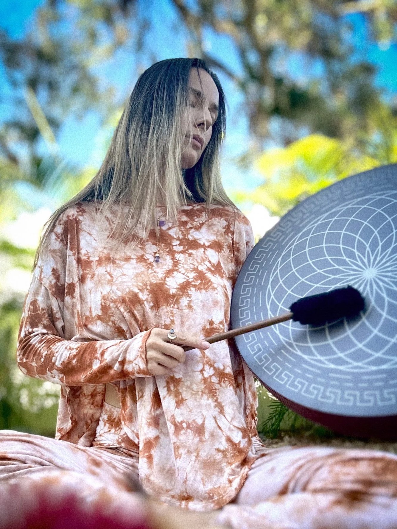 A woman sitting outdoors, playing a gong with a mallet, surrounded by trees and blue sky.