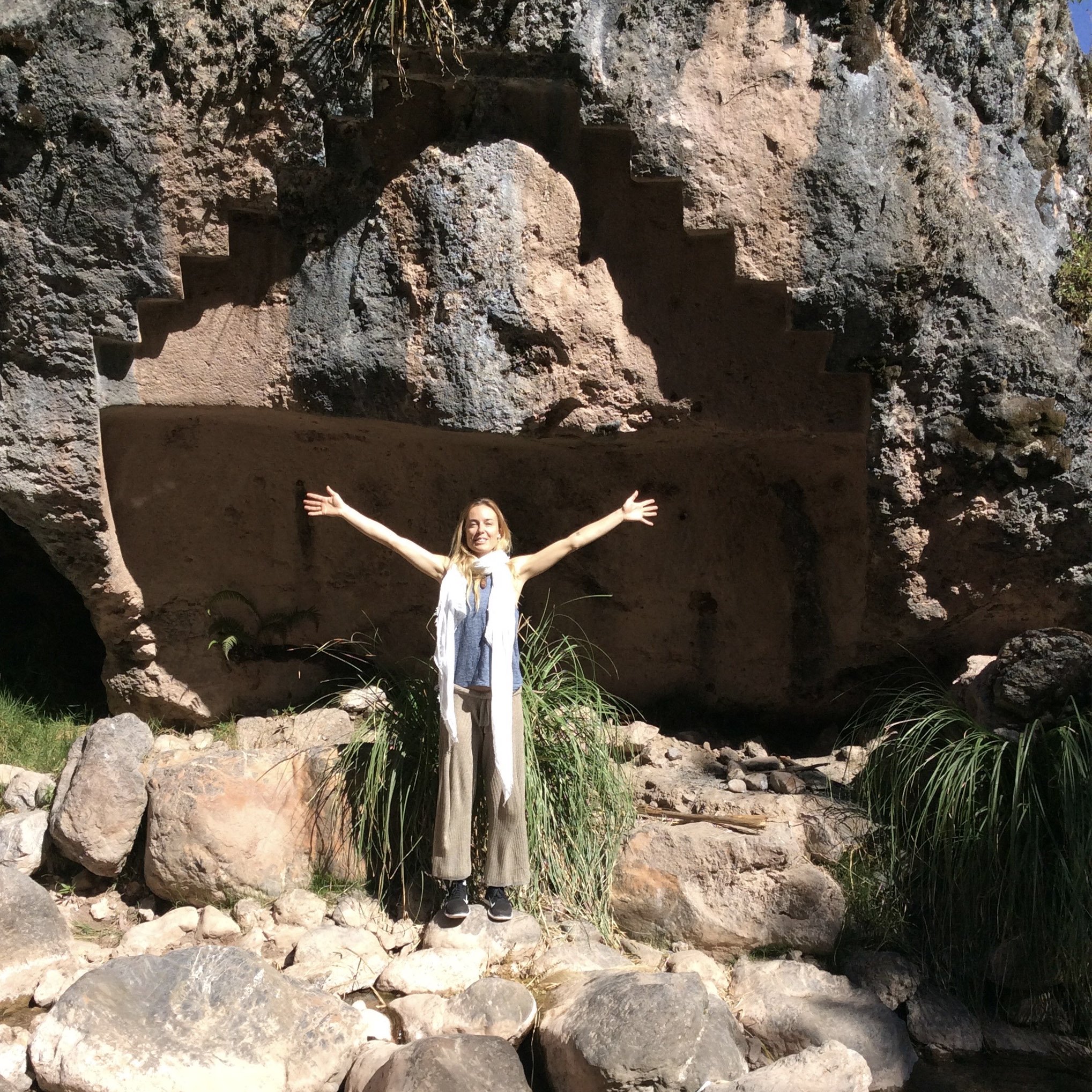 A woman with arms outstretched standing in front of a rock formation with carved stairs and a large face relief in an outdoor setting.