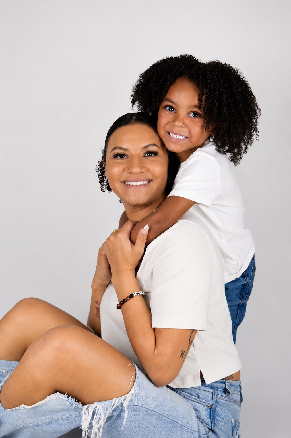 A woman with short dark hair and a young girl with curly hair smiling, with the girl on the woman's shoulders, both dressed in white shirts and blue jeans against a plain light gray background.