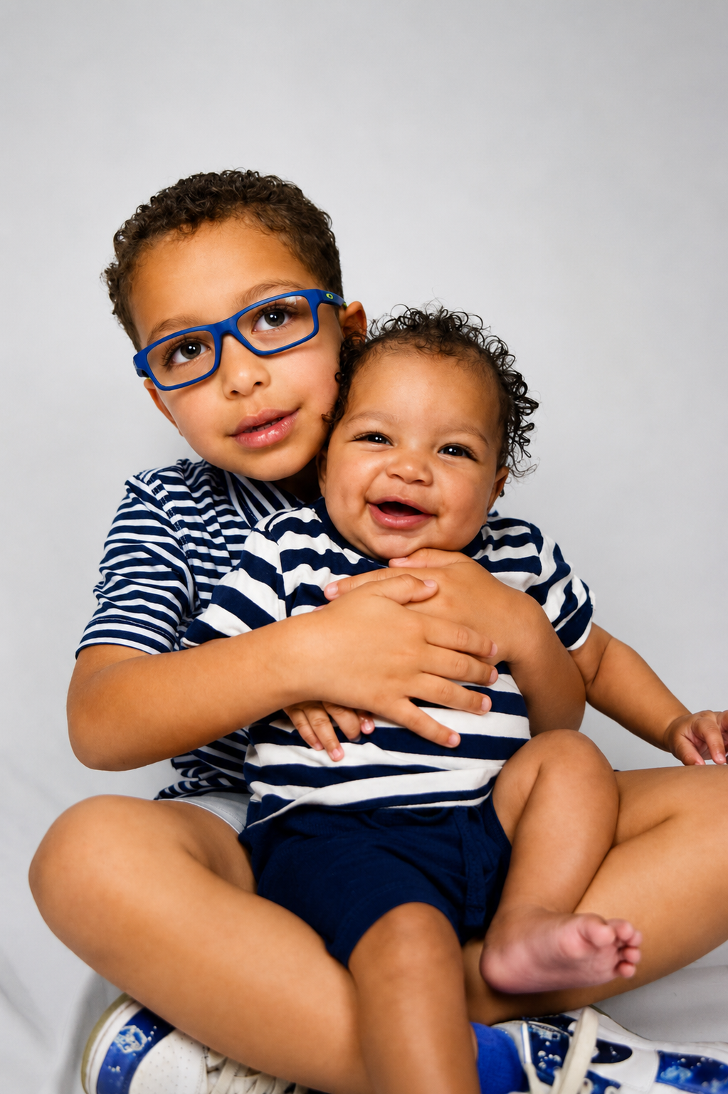 Two young children sitting on a white surface, a boy with glasses and a girl, both wearing striped shirts, smiling and hugging each other.