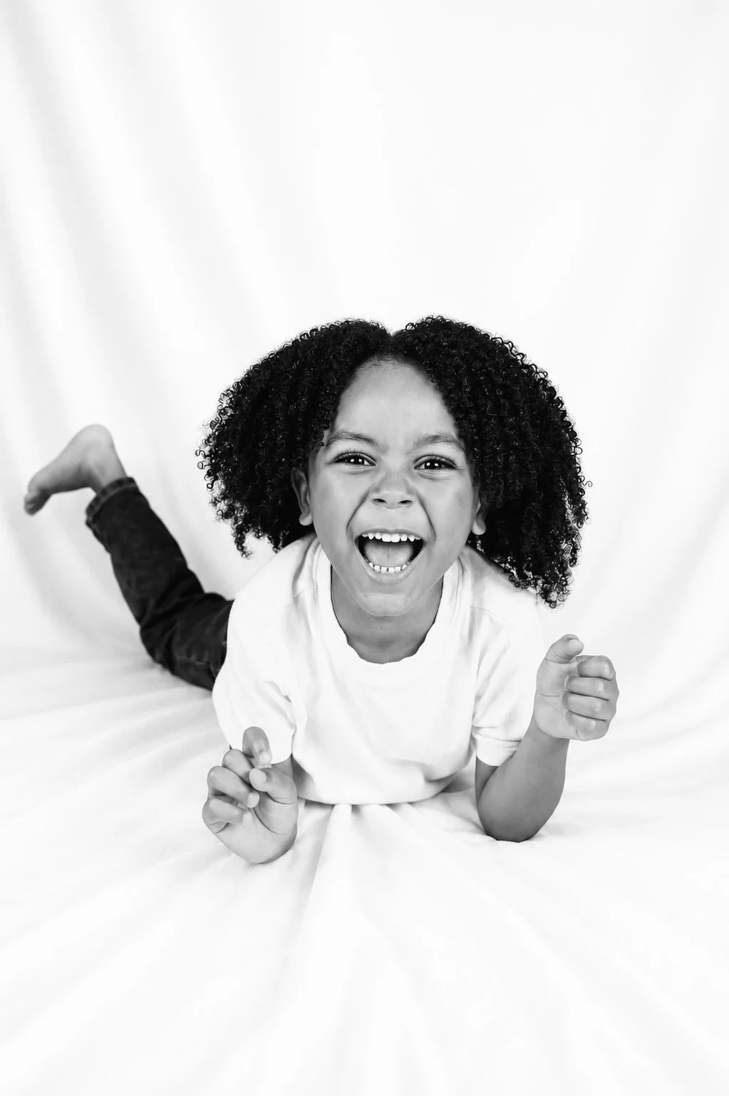 A young girl with curly hair lying on her stomach, smiling widely with her mouth open, wearing a white shirt and dark pants, against a plain light background.