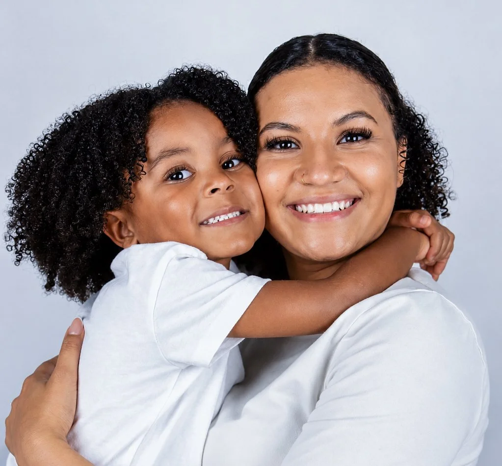 A happy woman and young girl hugging each other, both smiling, against a plain light gray background.