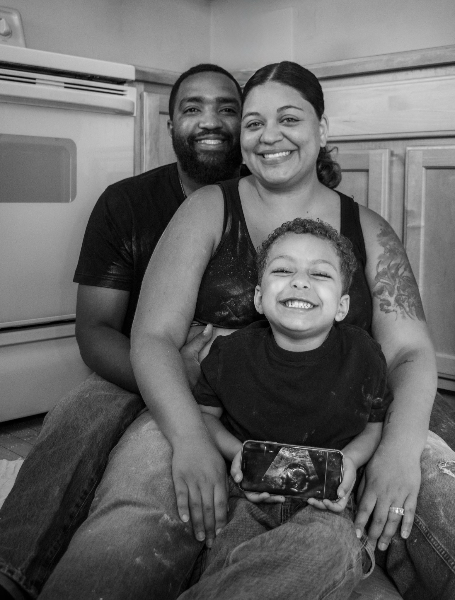 A smiling woman with a tattoo on her arm and a smiling young boy with curly hair, holding a phone with a picture of a whale. A man with a beard stands behind them, smiling, in a kitchen with wooden cabinets and an oven.