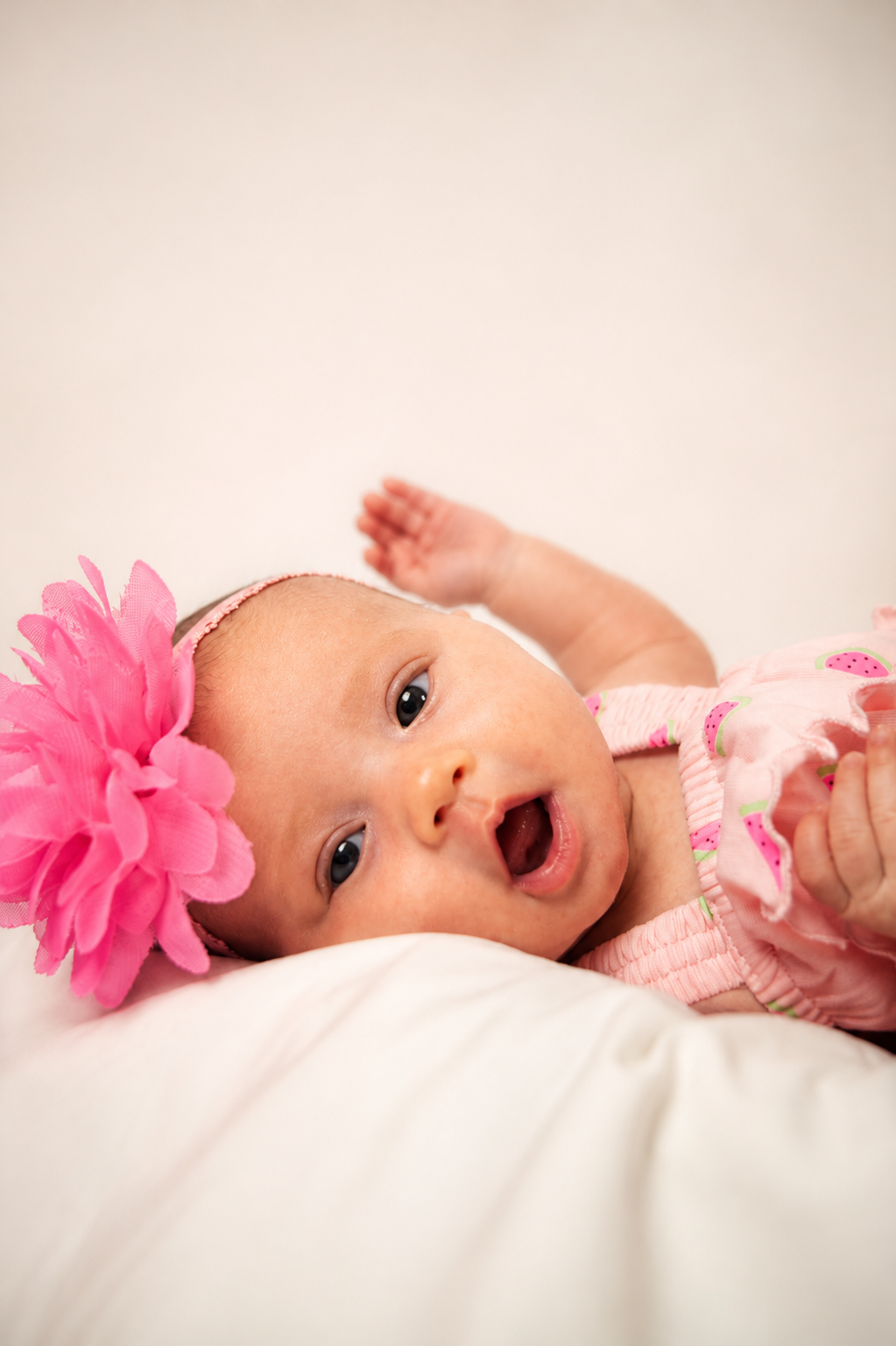 A baby girl lying on a white surface, wearing a pink dress with watermelon print and a large pink flower headband. She has her mouth open and looks curious.