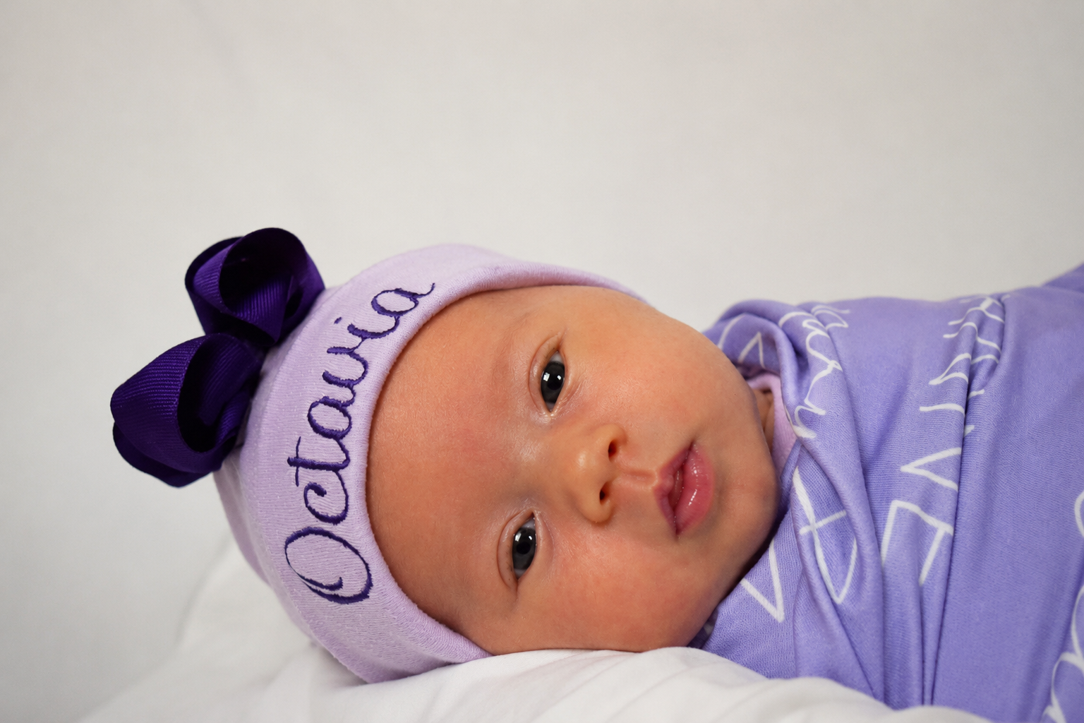 Close-up of a newborn baby with a lavender hat with a purple bow and the word 'Cutie' embroidered, lying on a white surface, dressed in a purple outfit.