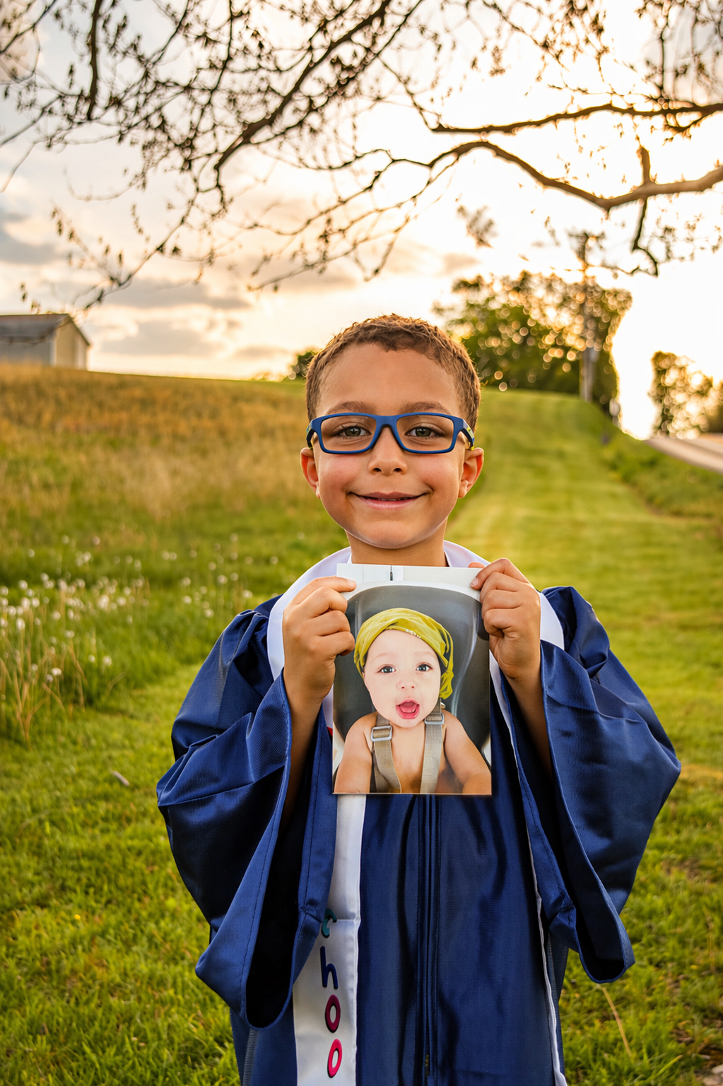 A young boy with brown hair, glasses, and a jacket, smiling outdoors at sunset, holding a photo of a younger child with a yellow headband.