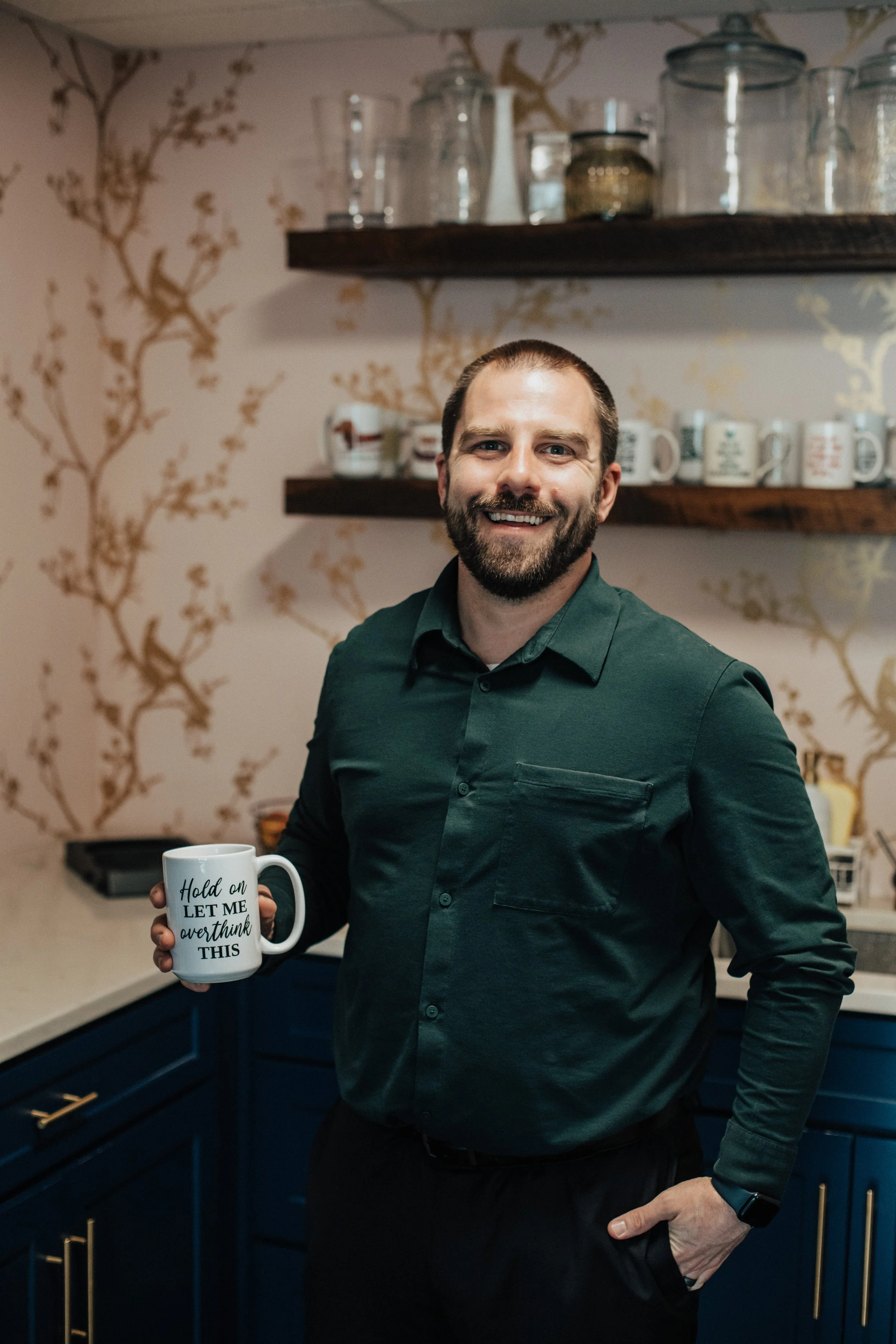 A man with a trimmed beard and short hair, wearing a green button-up shirt, smiling while holding a white mug with the words 'Hold on, let me overthink this' printed on it. He is standing in a kitchen with blue cabinets, a dark wooden shelf with glass jars and cups, and a decorative floral wallpaper background.