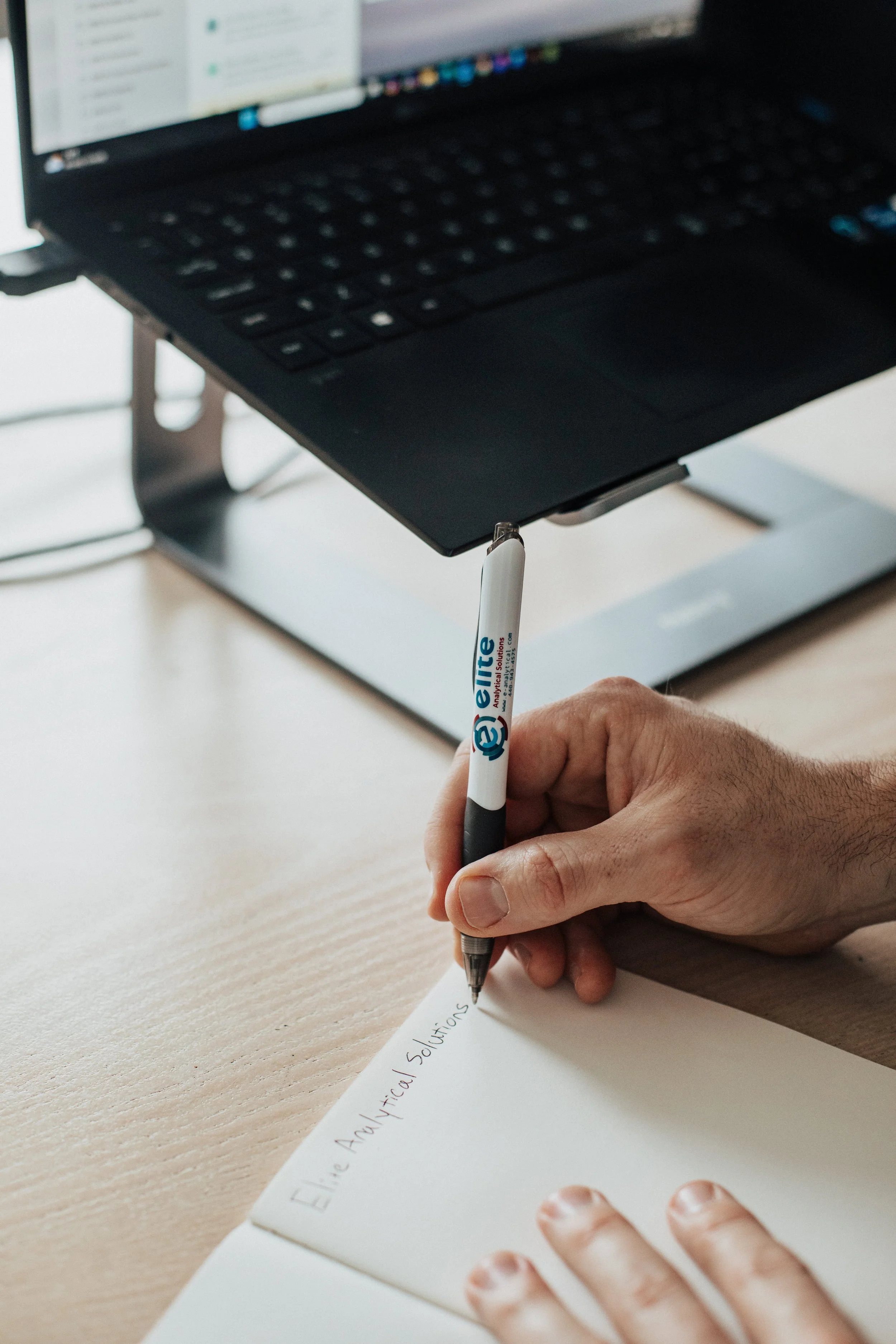 A person is writing in a notebook with a black and white pen, with the words 'Ene Analytical Solutions' written on it. The person's other hand is resting on the paper, and a laptop with a slightly blurred screen is visible in the background.