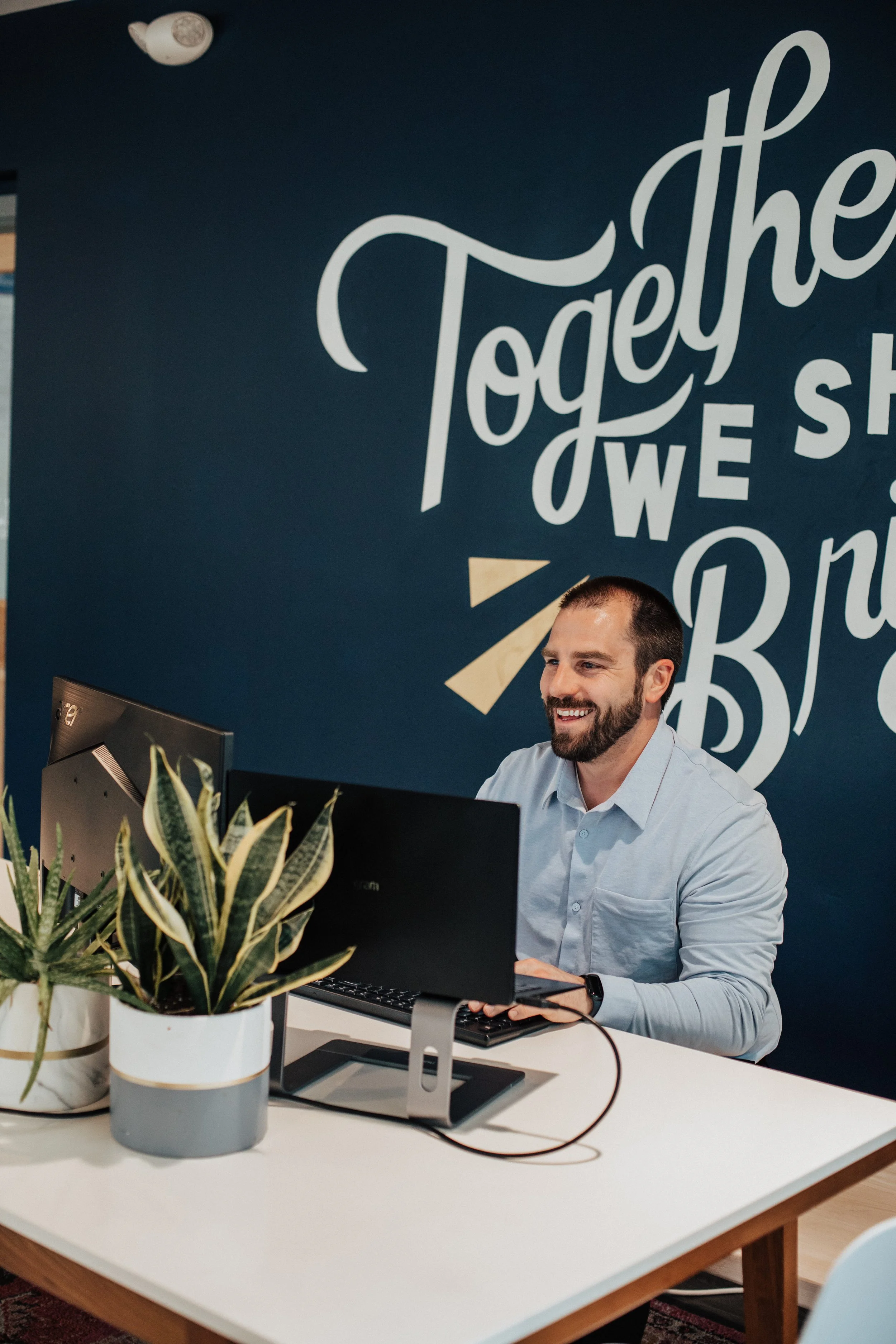 A man working at a desk with a computer, plants on the desk, and a large wall sign reading 'Togethe' in the background.