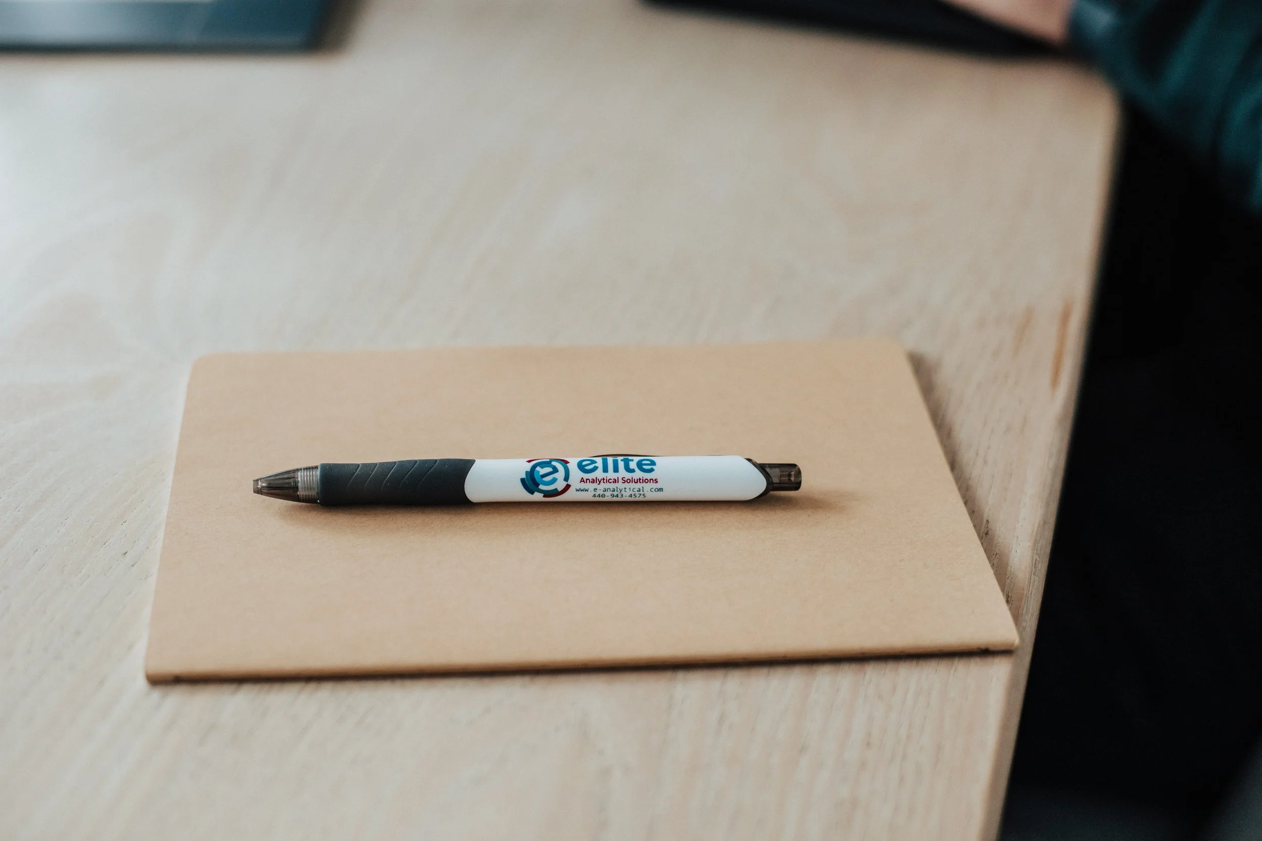 A black and white plastic ballpoint pen with the logo of Elite Analytical Solutions on a piece of beige paper on a wooden surface.