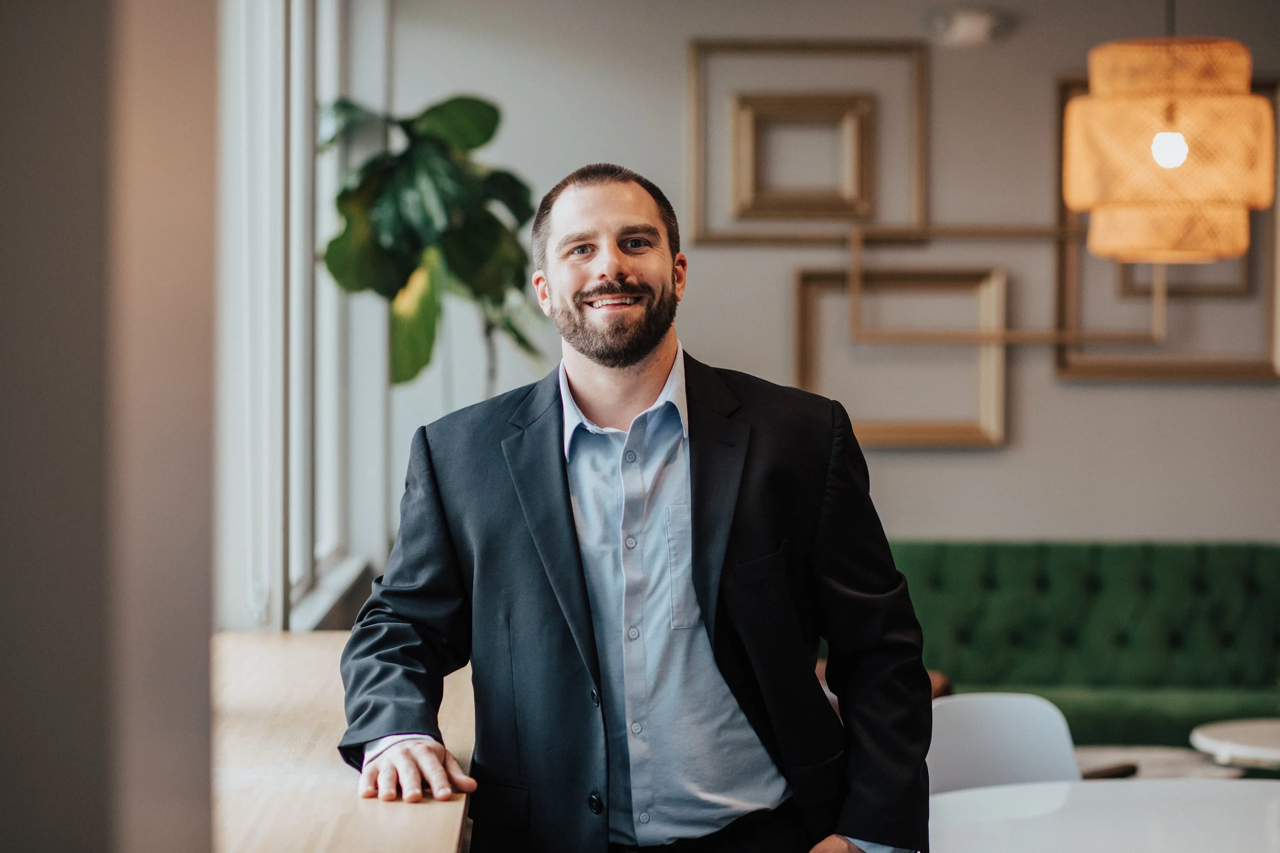 A smiling man with a beard, wearing a dark suit and light blue shirt, standing in a modern office or conference room with wooden frames on the wall and a hanging woven light fixture.