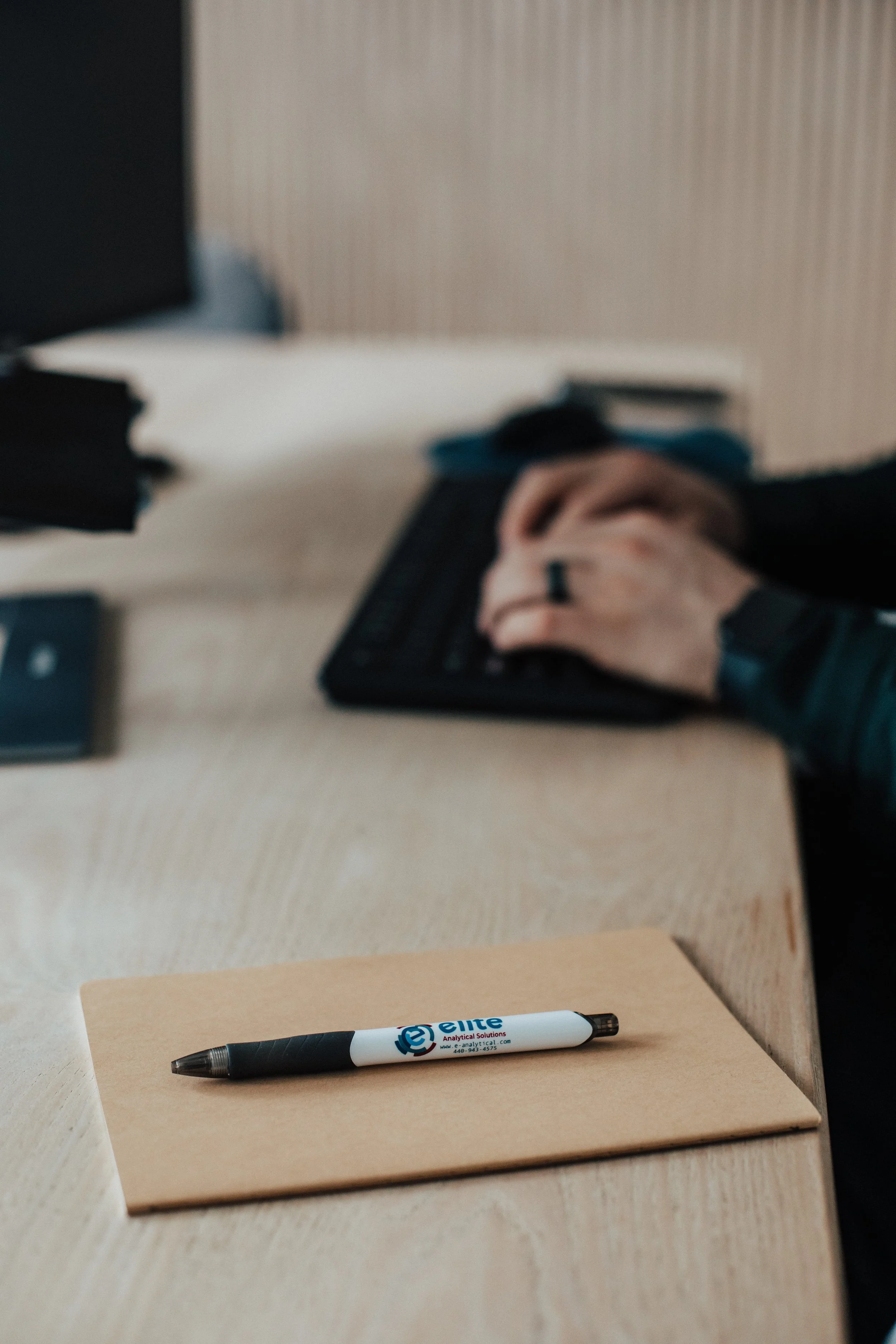 Close-up of a black and white marker pen on a beige notebook placed on a wooden table, with a blurred background including a person's hand, a computer keyboard, and a monitor.