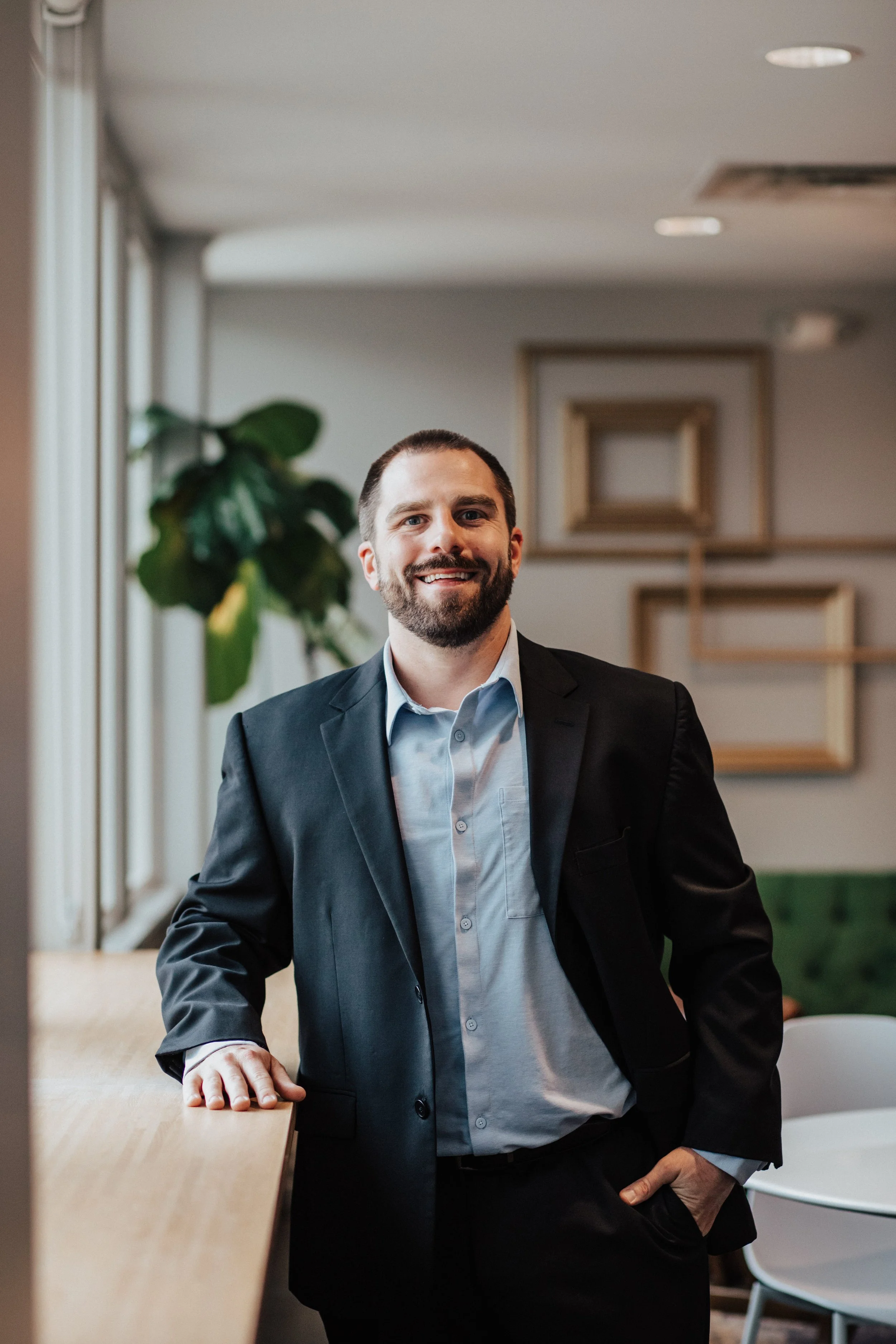 A man with a beard in a suit smiling while leaning on a wooden table in a modern office or conference room with framed art on the wall and a plant.