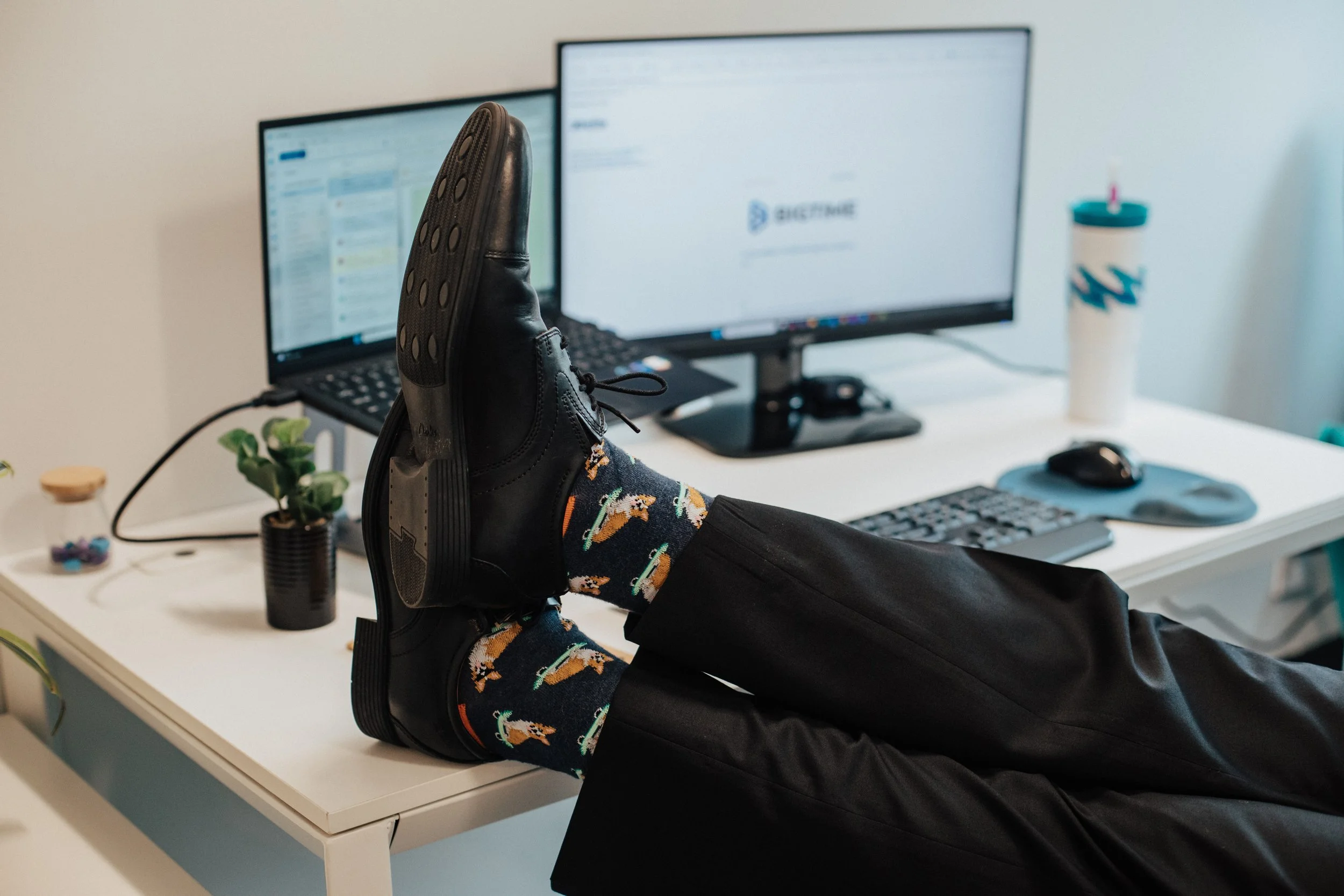 Person resting their feet on a white desk, wearing colorful socks with horses and black shoes, in an office with dual monitors, plants, and a tumbler.