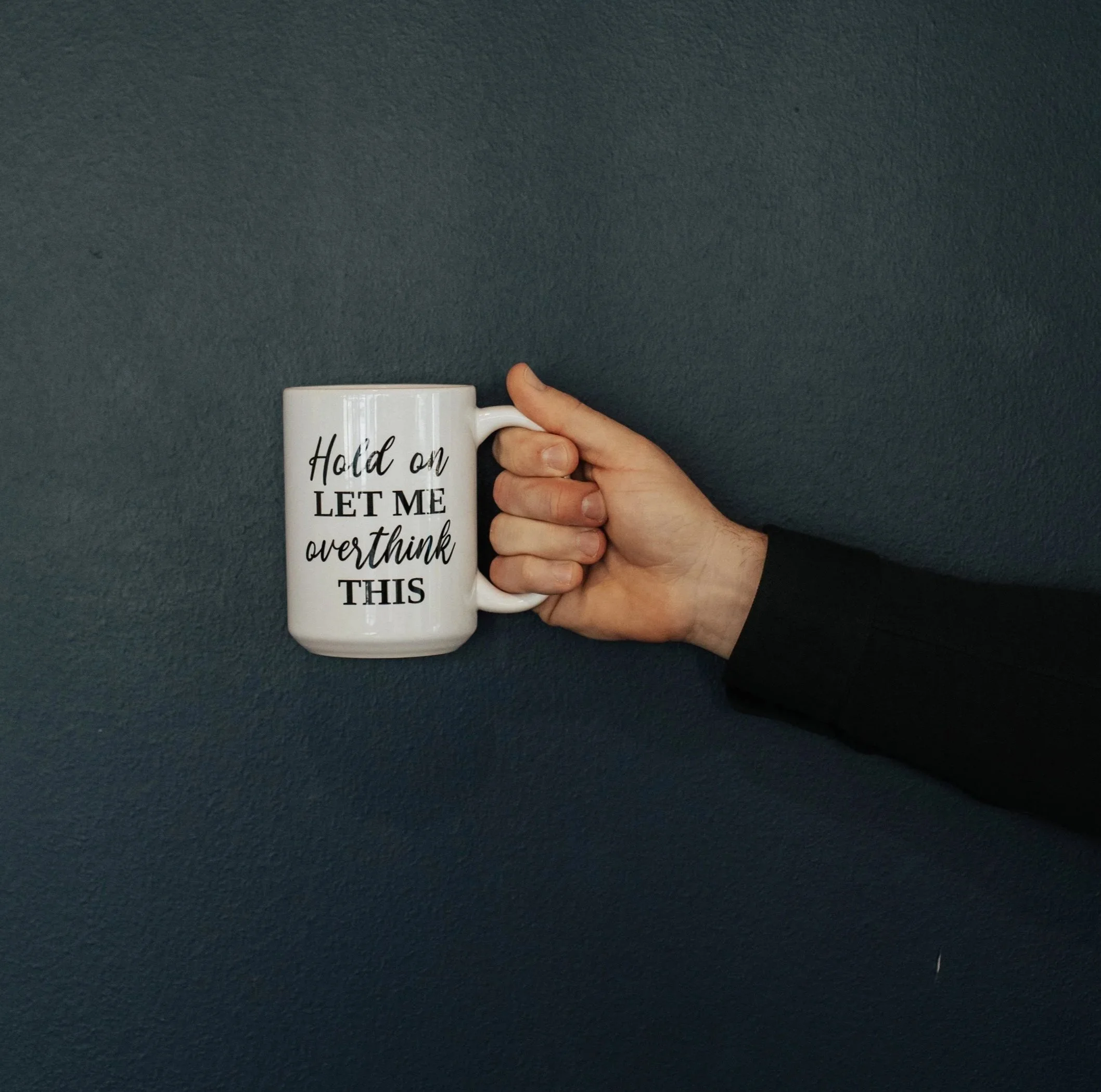 A person's hand holding a white coffee mug with black text against a dark textured background.