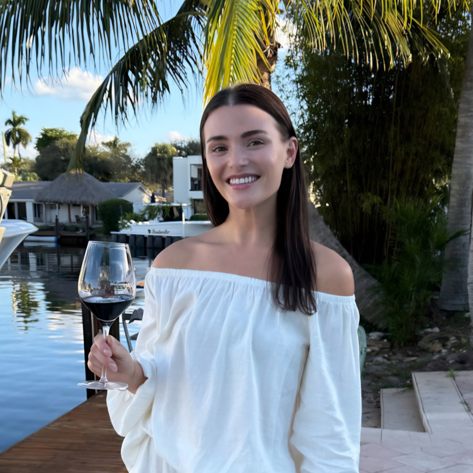 Smiling woman holding a glass of red wine outdoors with palm trees, a dock, and water in the background.