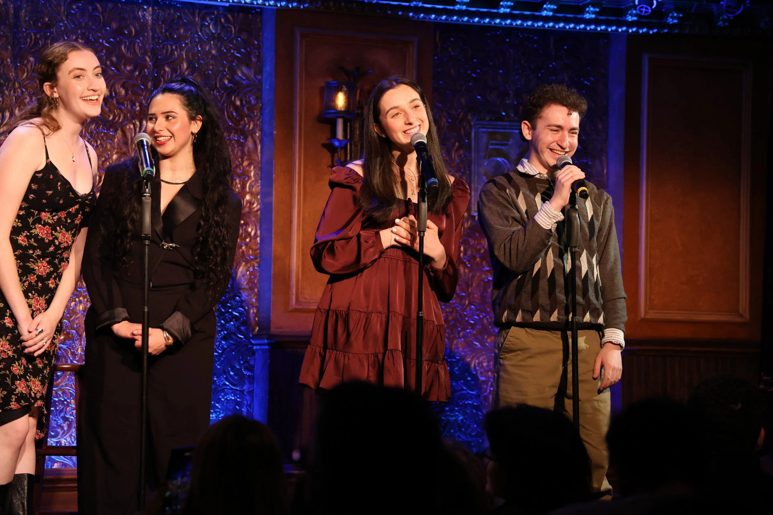 Four young adults standing on stage with microphones, smiling and engaging in a performance or presentation, with an ornate background and warm lighting.
