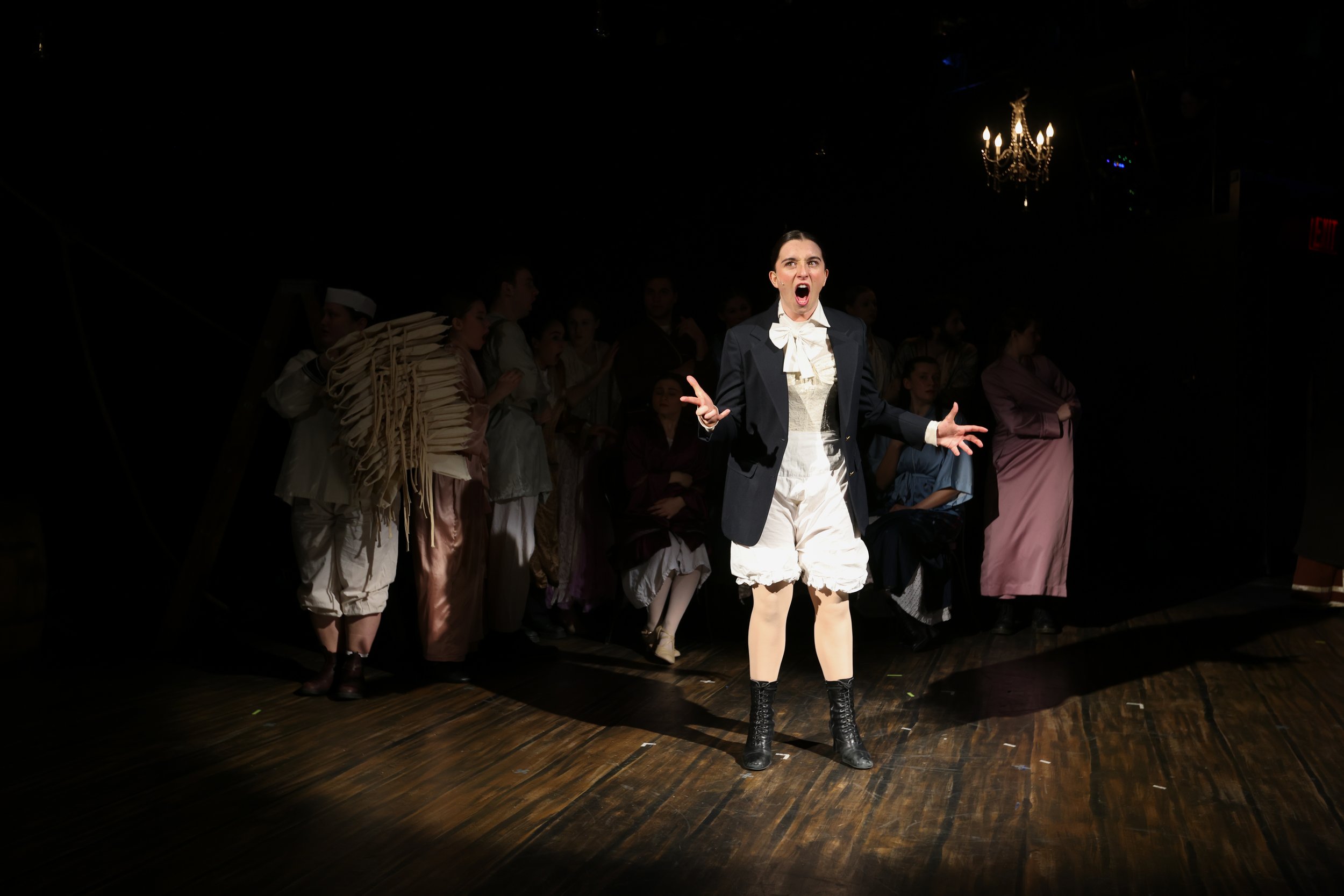 A woman with dark hair in a costume, likely from a play, standing on a stage with a group of people dressed in period costumes behind her. The woman appears to be shouting or singing passionately, with a chandelier hanging above.