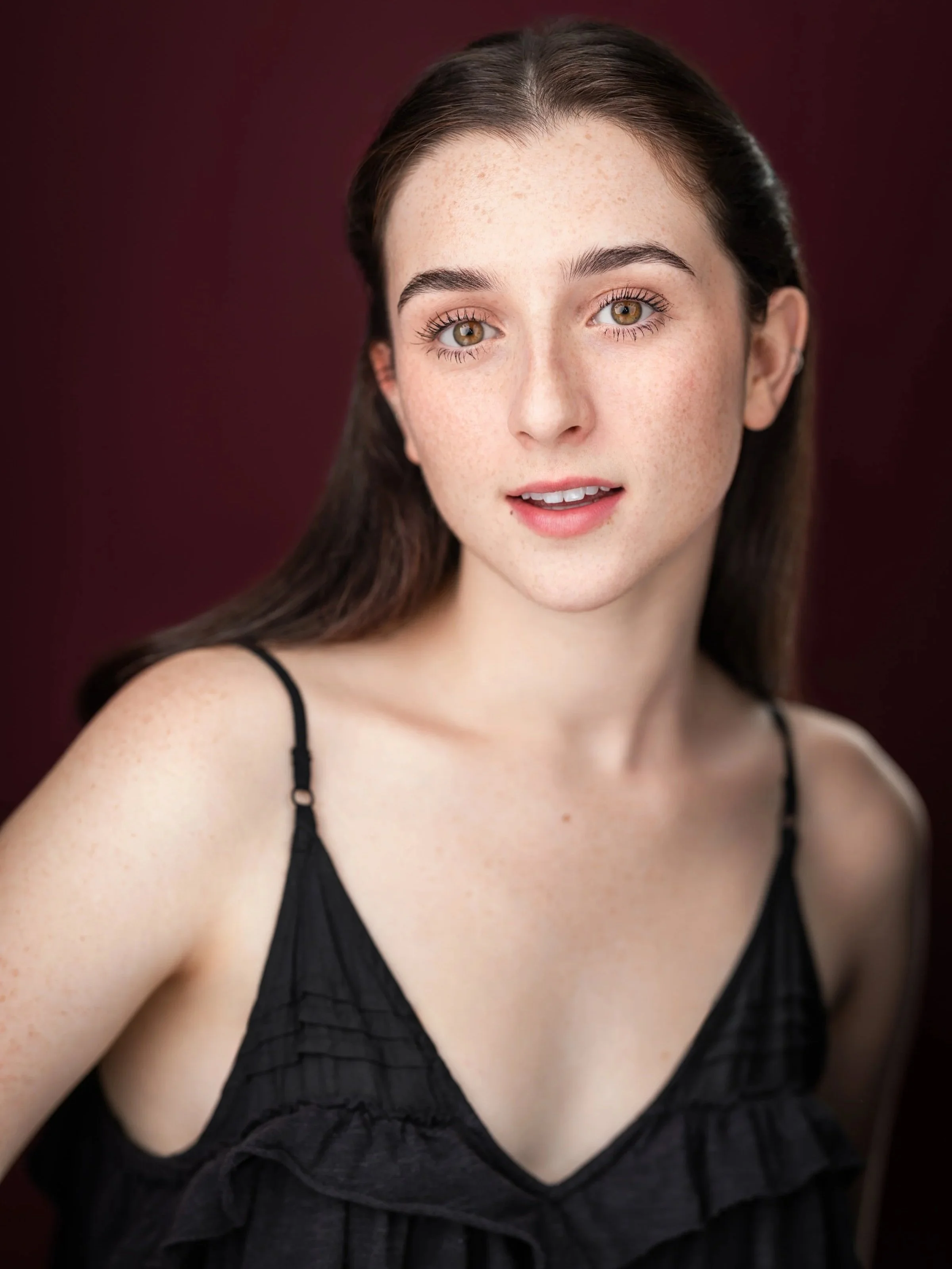 Close-up of a young woman with brown hair, hazel eyes, and freckles, wearing a black spaghetti strap top, posing against a dark red background.