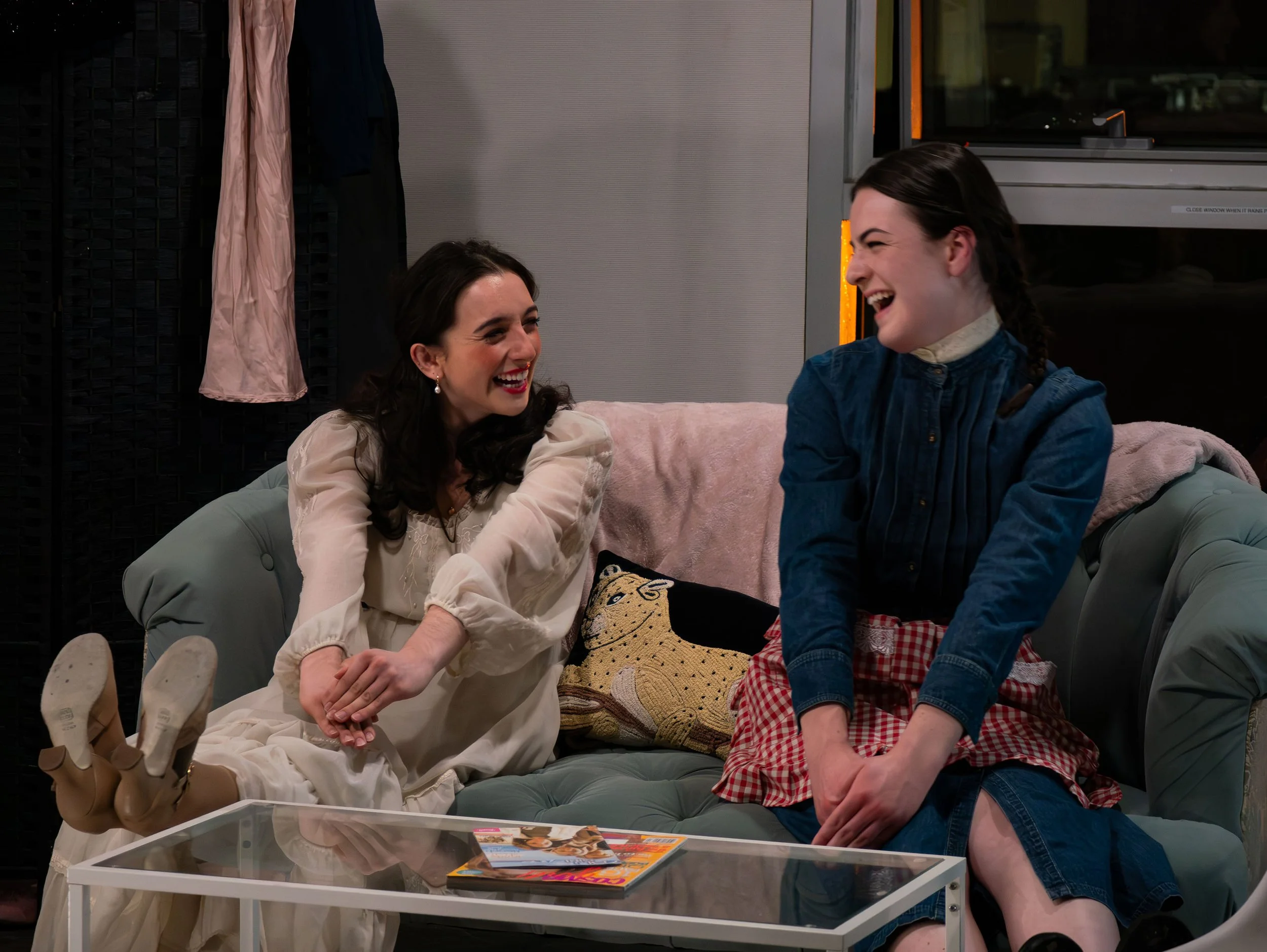 Two women laughing and talking while sitting on a green tufted sofa in a modern living room. One woman is dressed in a white dress and the other in a blue shirt and red checkered skirt. There is a glass coffee table with magazines in front of them an