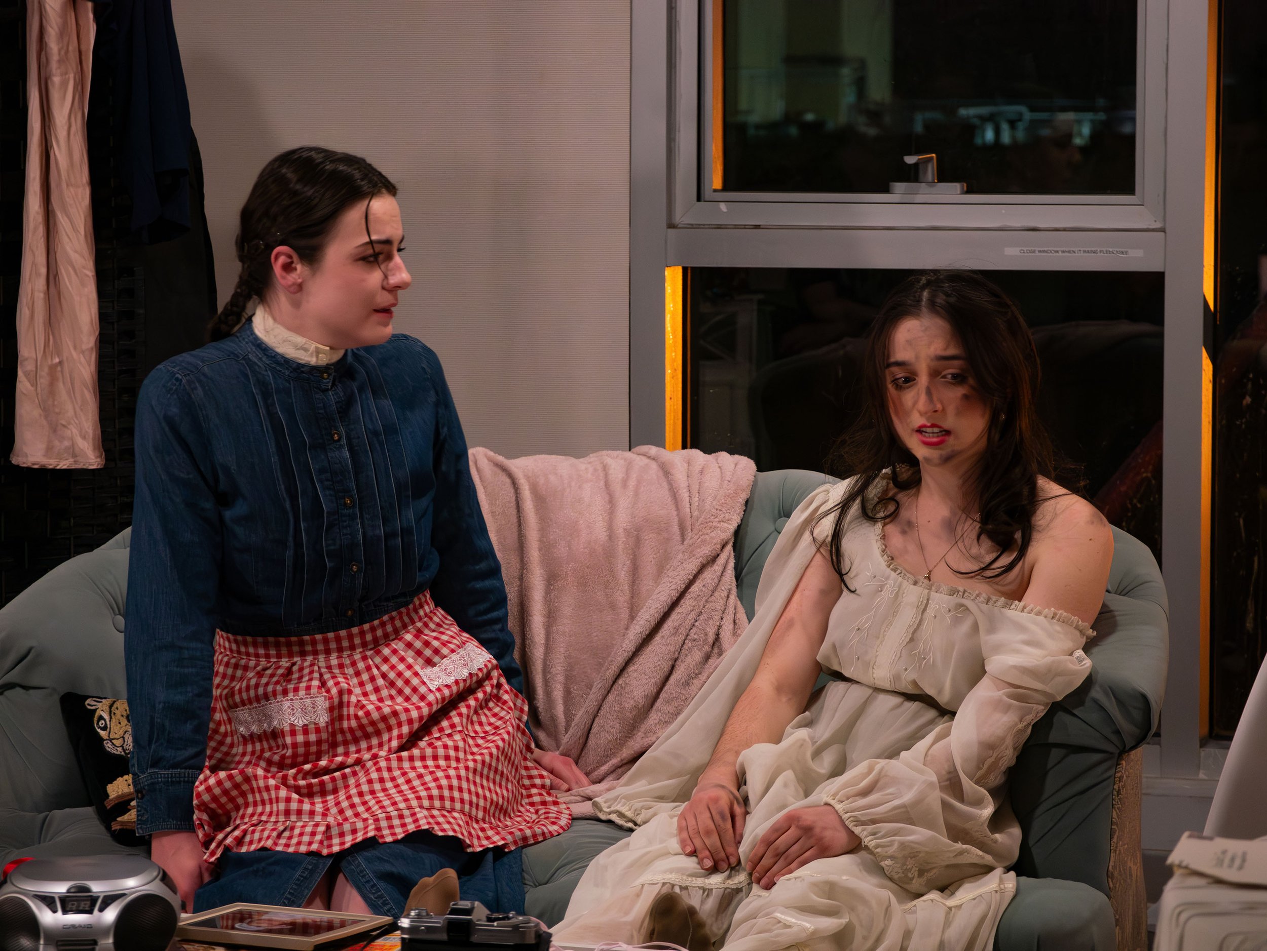 Two women in a living room, one sitting on a couch wearing a nightgown, the other standing and wearing pajamas, having a conversation with distressed expressions.