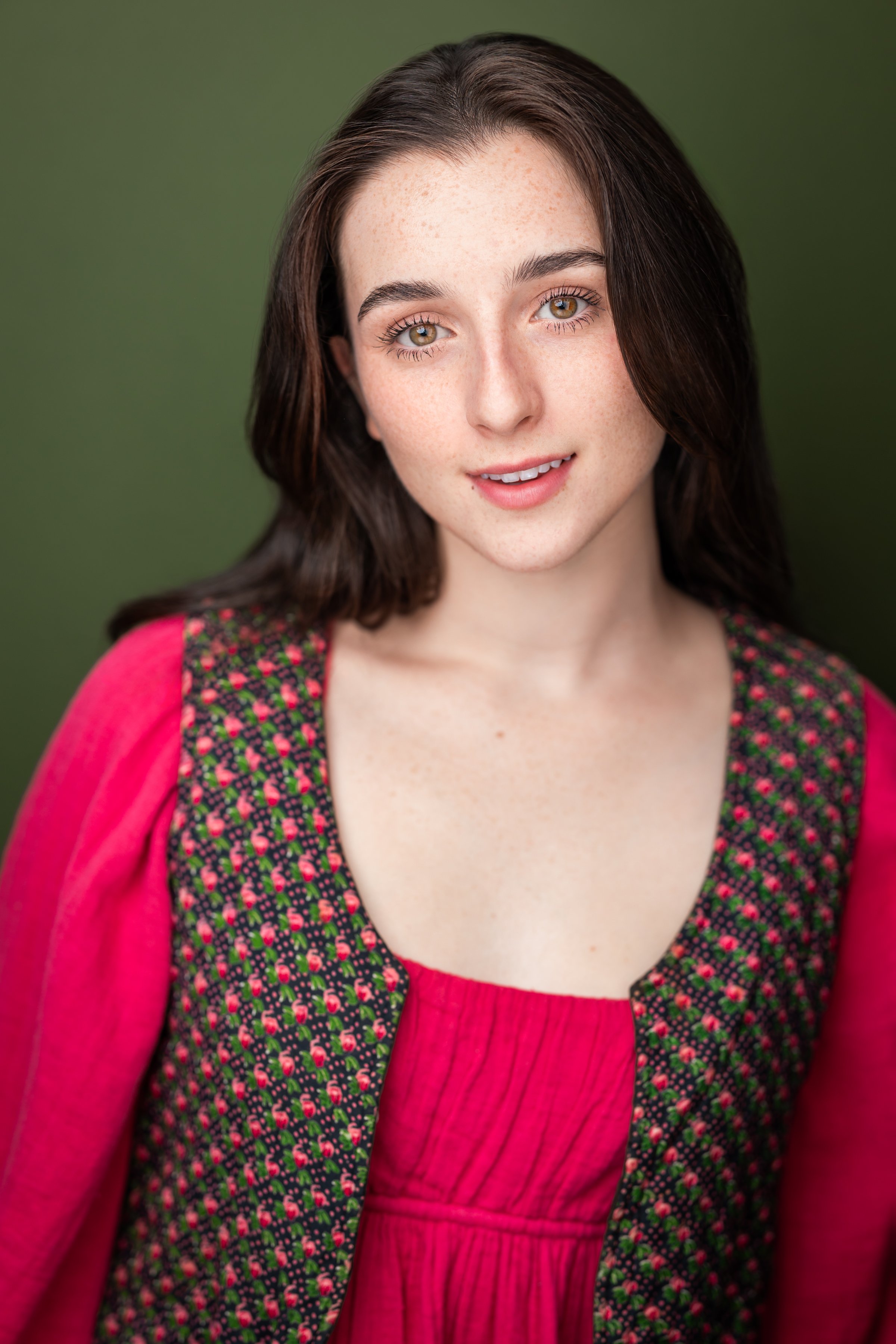 Close-up of a young woman with long dark hair, hazel eyes, and freckles, wearing a red dress and a colorful floral jacket, smiling softly against a green background.
