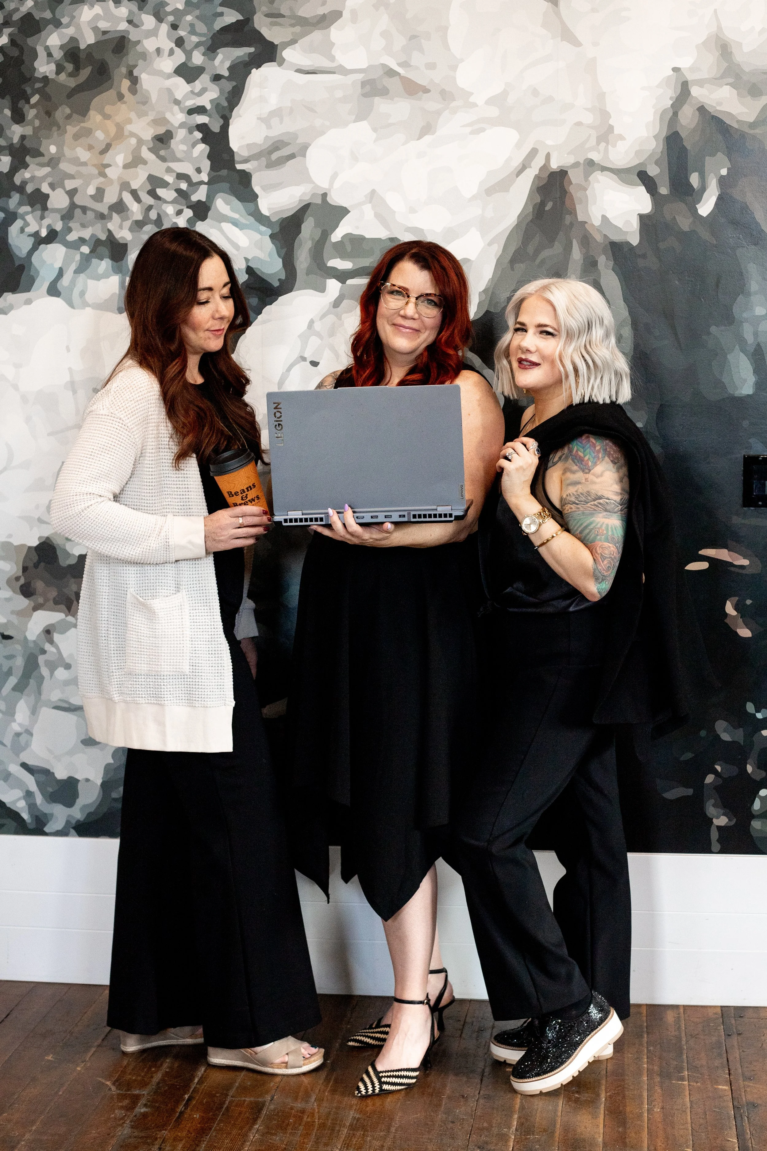 Three women standing together, one holding a laptop, in front of a large floral mural wall.