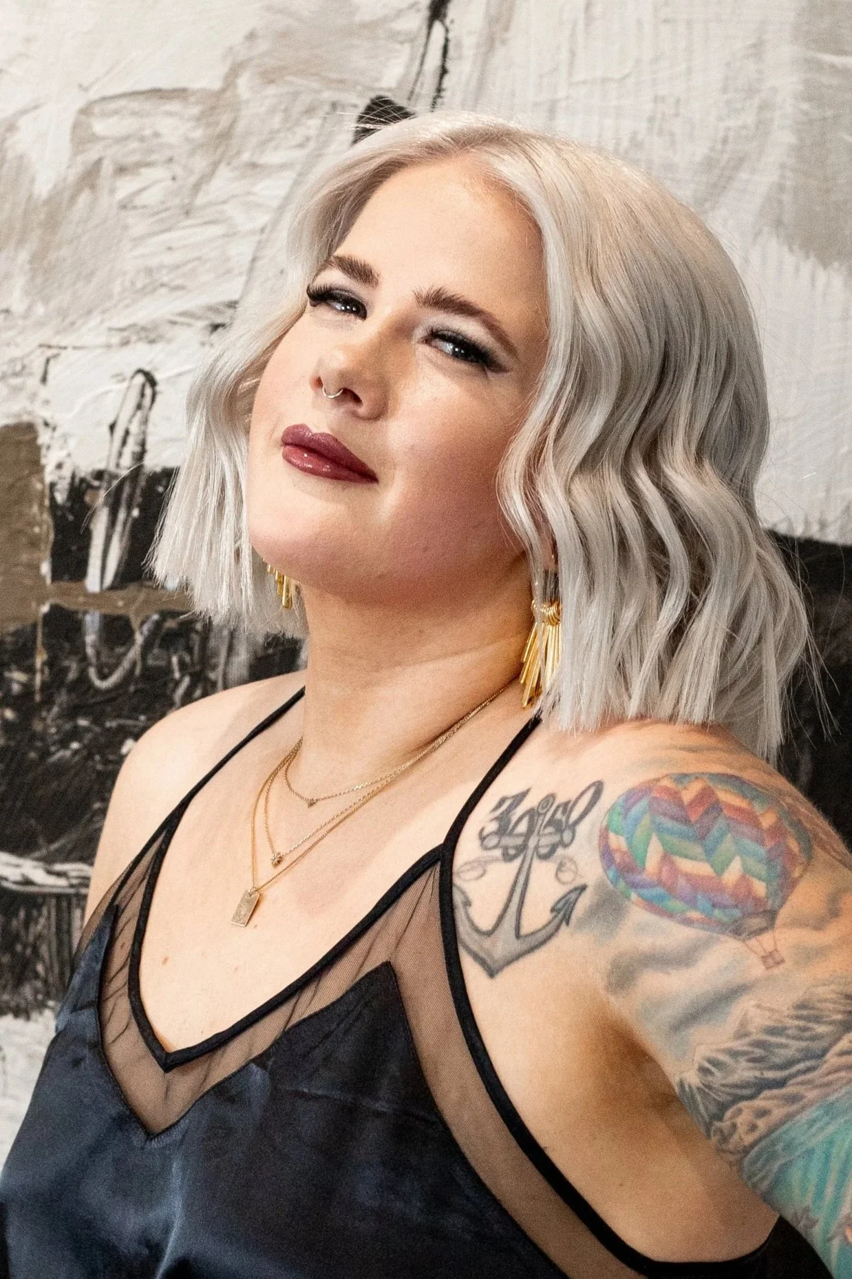 A woman with platinum blonde wavy hair, tattoos on her arm, wearing gold earrings, layered necklaces, and a black sheer top, smiling slightly in front of a black and white background.