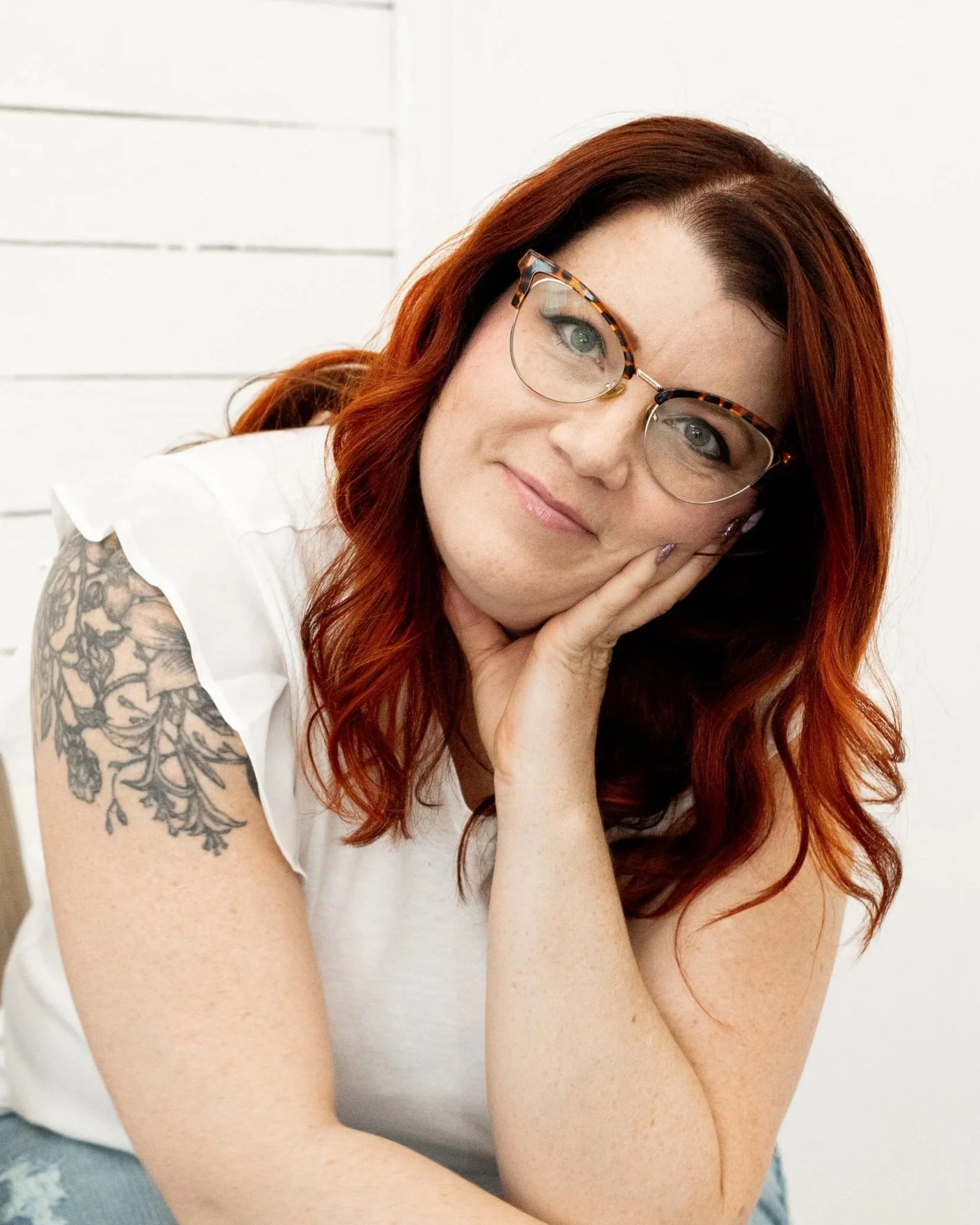 Close-up of a woman with red hair, glasses with tortoiseshell frames, and a tattoo on her upper arm, resting her chin on her hand with a gentle expression. She is wearing a white top and appears to be indoors with a white wall in the background.
