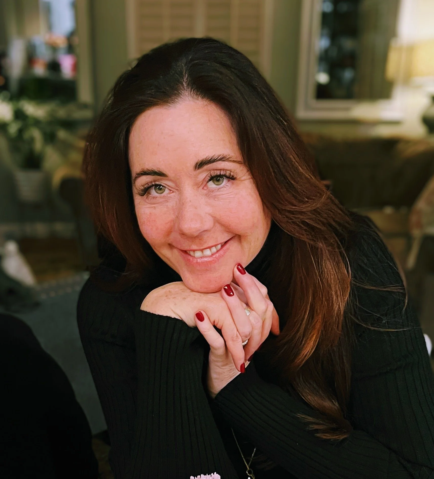 A woman with long brown hair and green eyes, smiling, resting her chin on her hands, wearing a black sweater with red nail polish, in a cozy indoor setting.