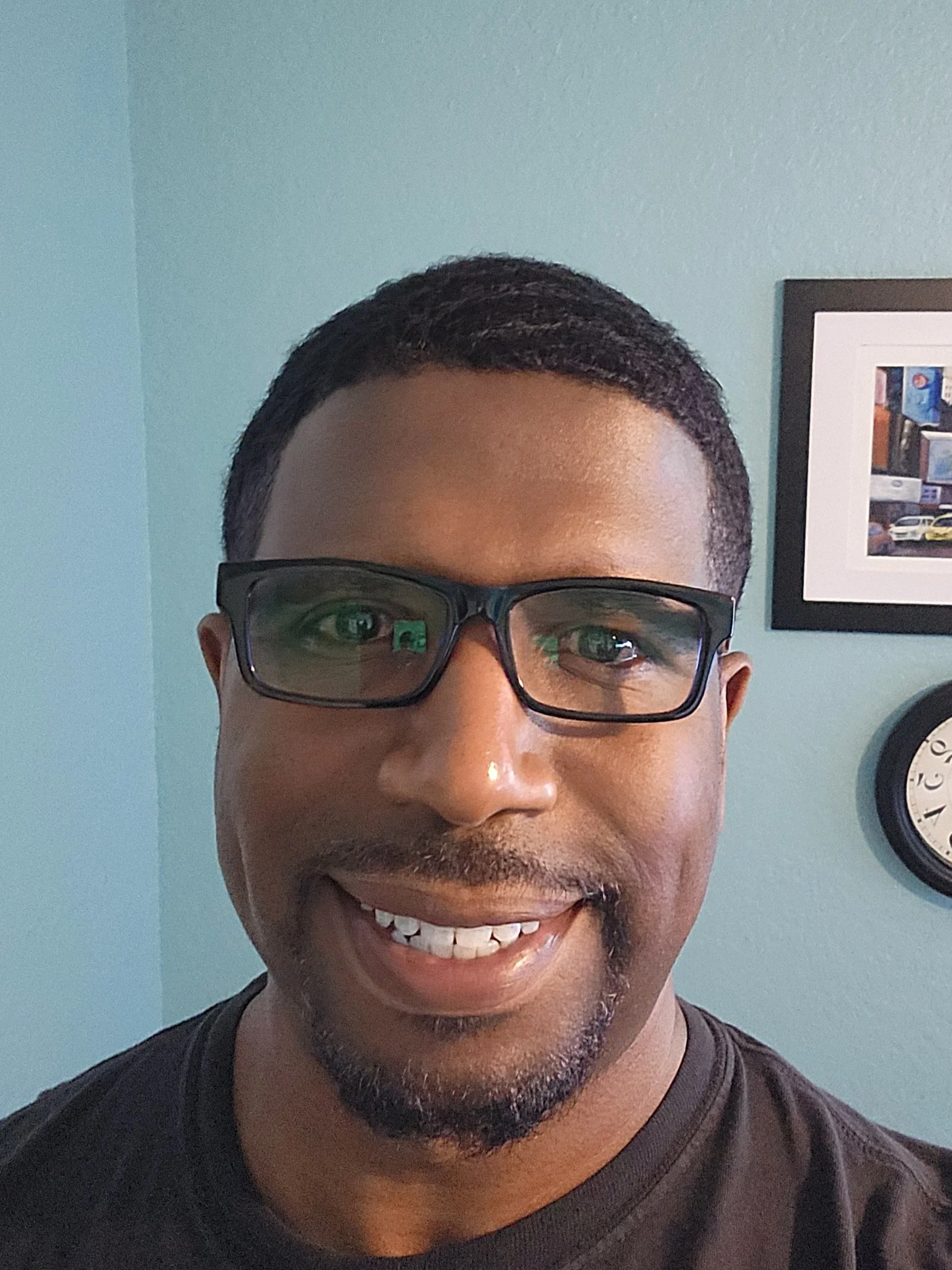 A man with glasses smiling for a photo indoors, with a framed picture and a clock on the wall behind him.