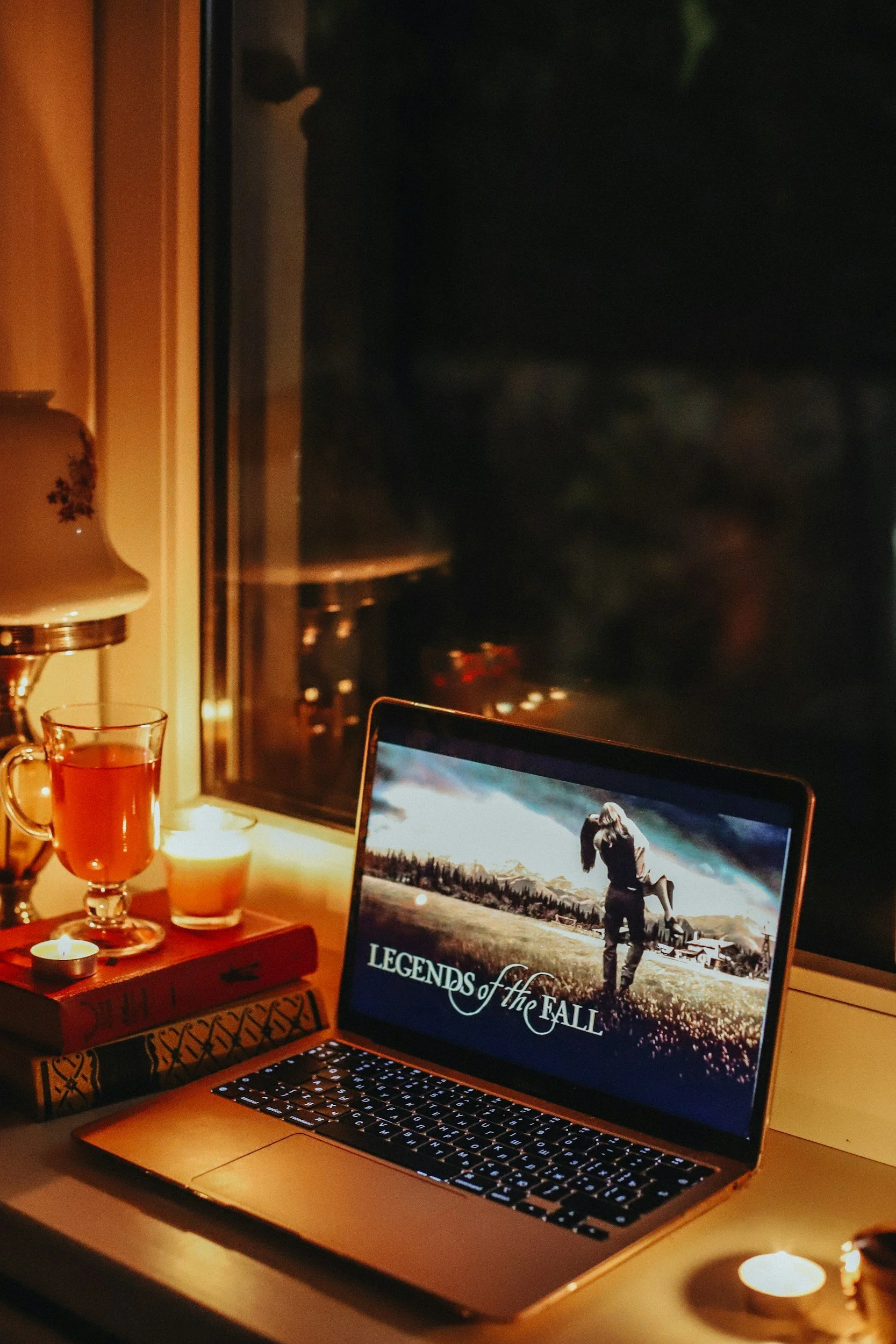 A laptop on a desk displaying the title "Legends of the Fall" with a scenic background. Nearby are lit candles, a glass cup with a warm beverage, and a few books. The scene is set at night, visible through a window.