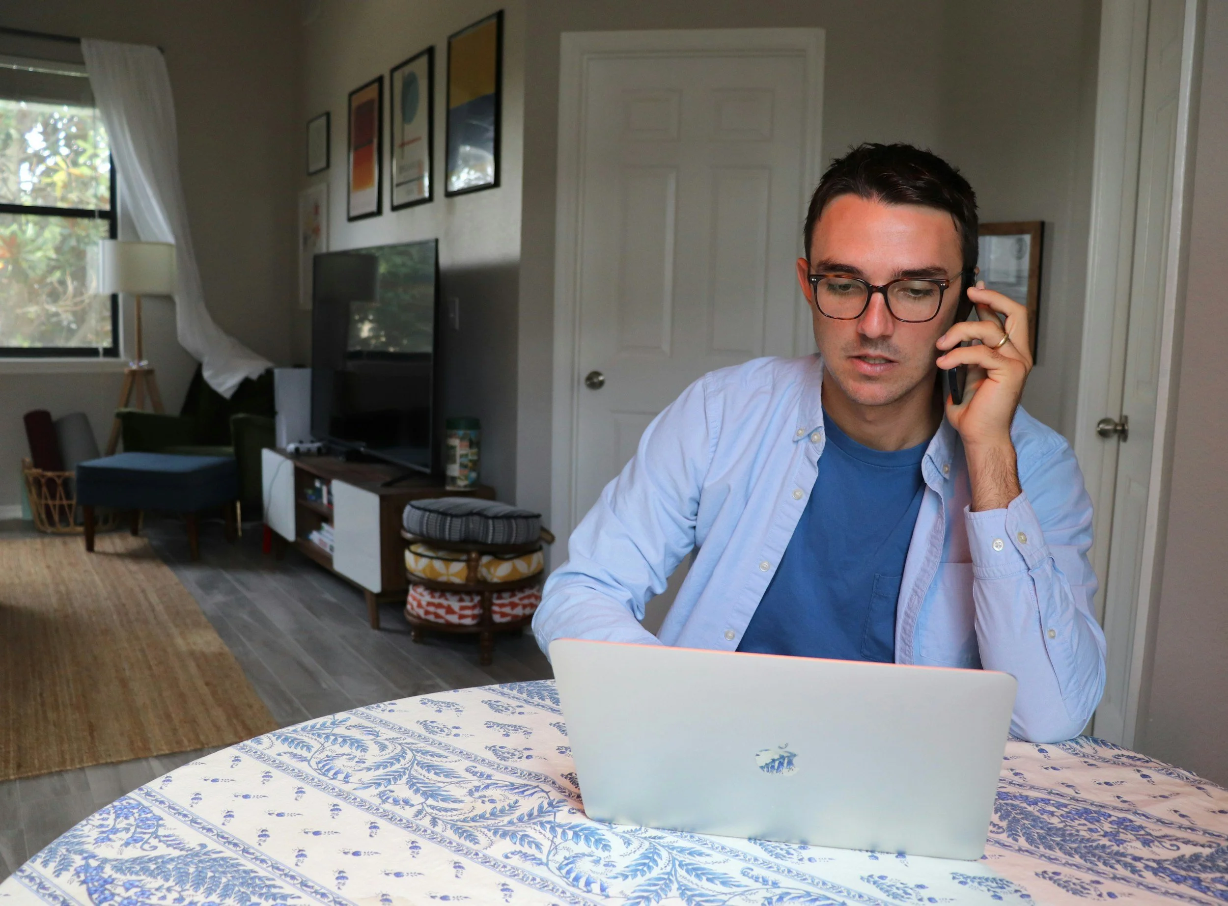 A man with glasses sitting at a table using a laptop and talking on a phone in a living room with a TV, artwork, and window in the background.