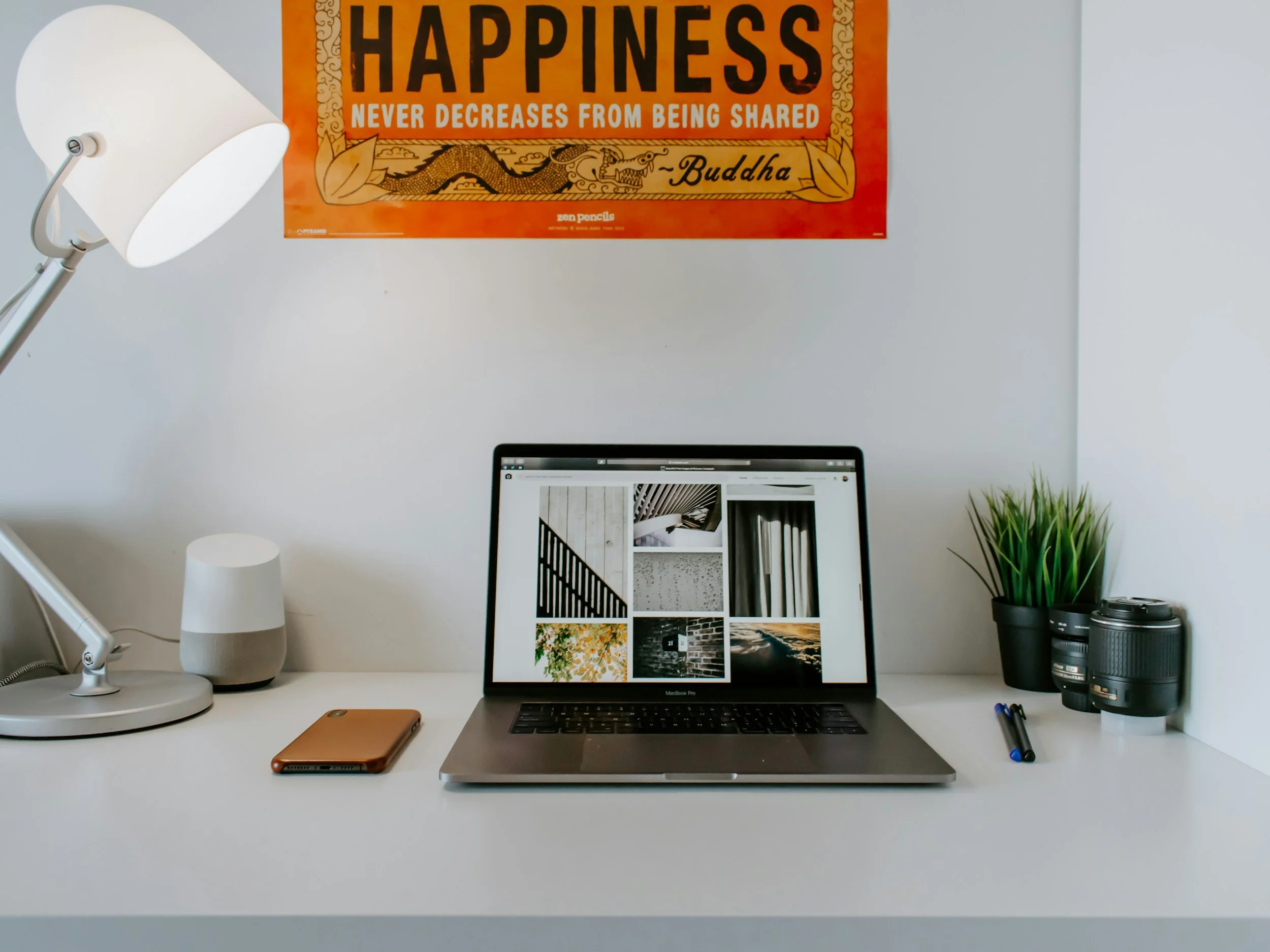 Minimalist white desk with a laptop displaying photo thumbnails, a brown notebook, two black pens, a potted green plant, two camera lenses, a white smart speaker, and a white desk lamp. On the wall behind, there is an orange poster with the quote 'Happiness never decreases from being shared' attributed to Buddha.