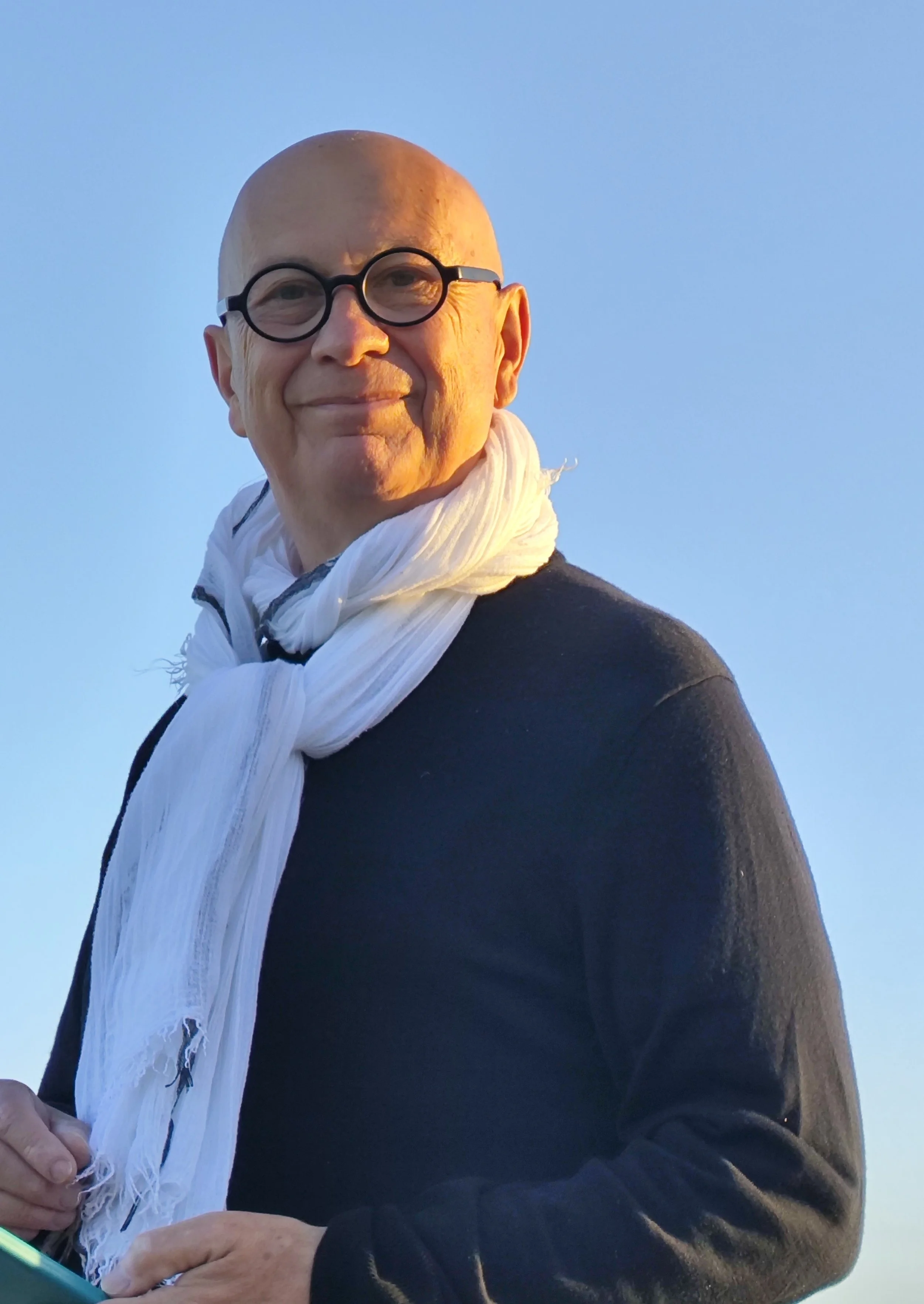 Un homme chauve portant des lunettes rondes noires, un pull noir et un foulard blanc, souriant, avec un ciel bleu en arrière-plan.