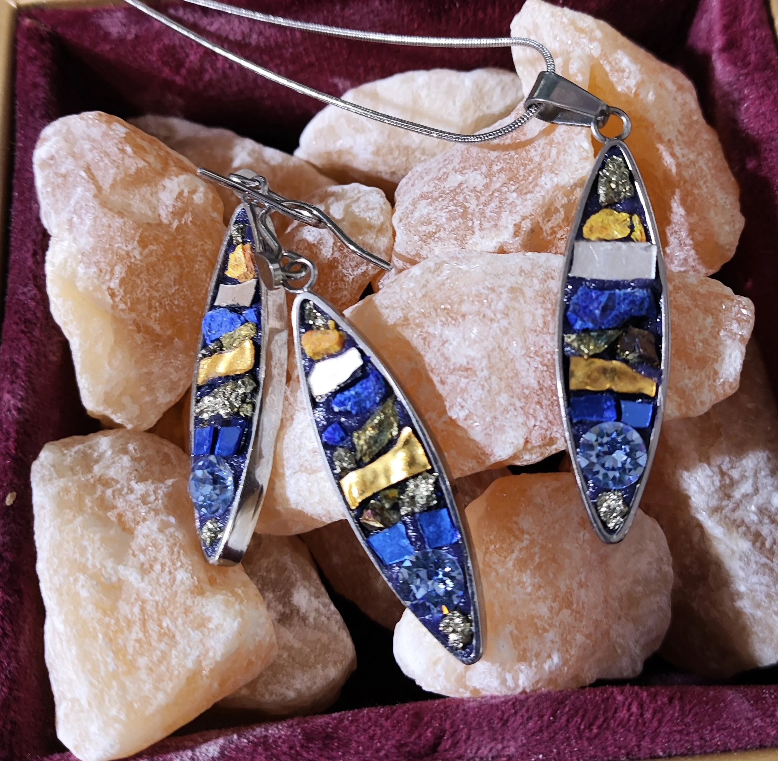 Collier et boucles d'oreilles avec des pierres en veine de couleur or, bleu, blanc et noir posés sur des morceaux de roche ou pierre en background rouge foncé.