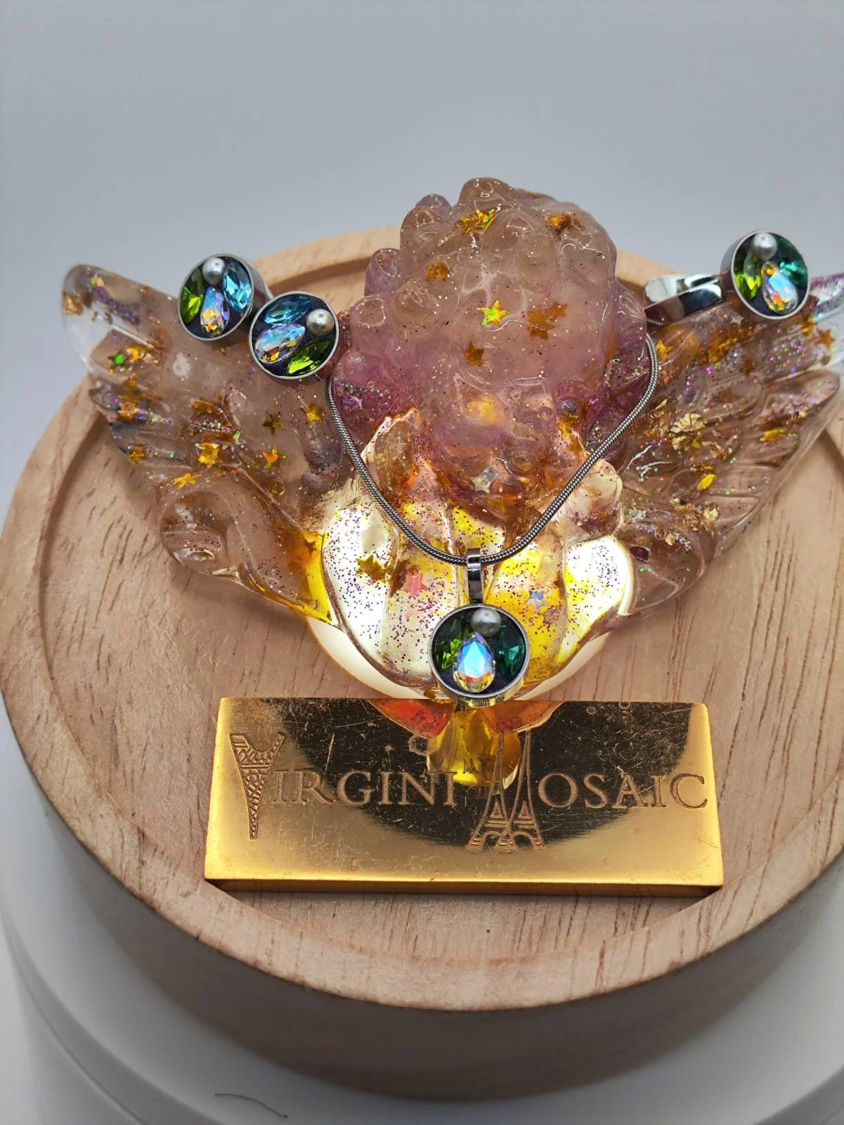 Bijoux en verre avec pendentif orné de pierres précieuses colorées, présenté sur un socle en bois avec plaque gravée portant la marque "Virgin Mosaic".