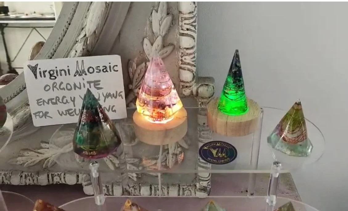 Cristaux en forme de pyramide de différentes couleurs sur un support en verre, avec une note écrite à la main indiquant 'Virgini Mosaic, Orgonite, Energy, Young, For Well Being'.